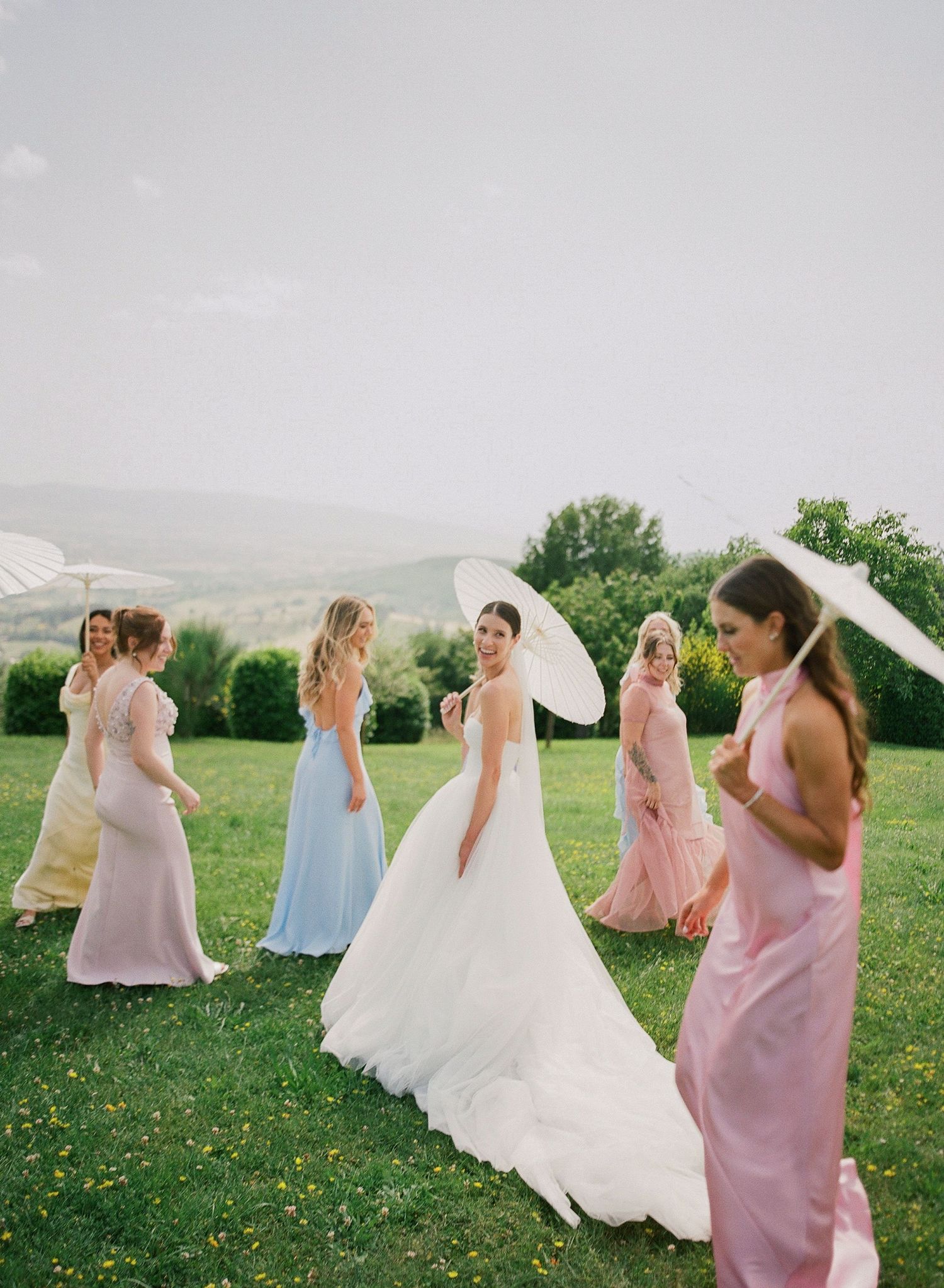 A bridal party in pastel bridesmaid dresses and flowing white wedding gown walk together on a grassy hilltop with mountains behind.