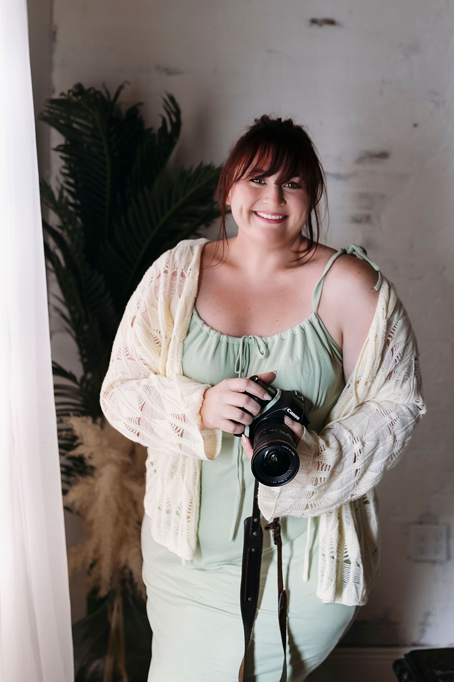 Smiling female photographer holding a DSLR camera, standing in a naturally lit studio with tropical plants, embodying pr
