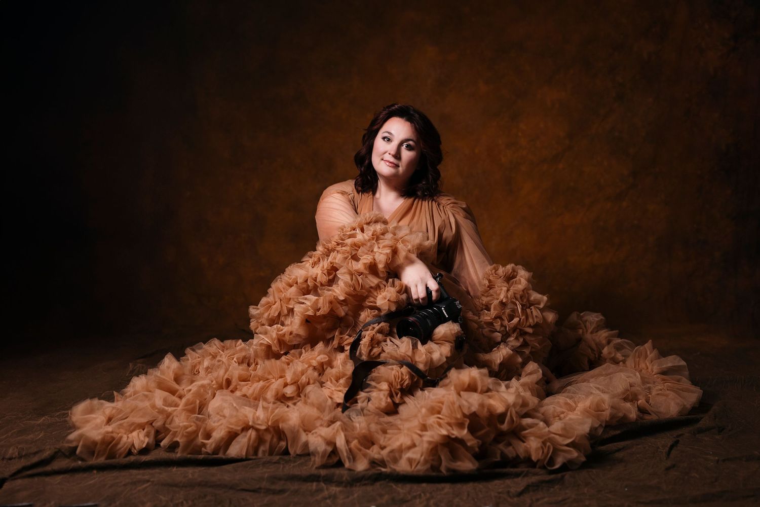 Portrait photographer in a voluminous tulle dress holding a camera, with a warm, artistic studio backdrop.