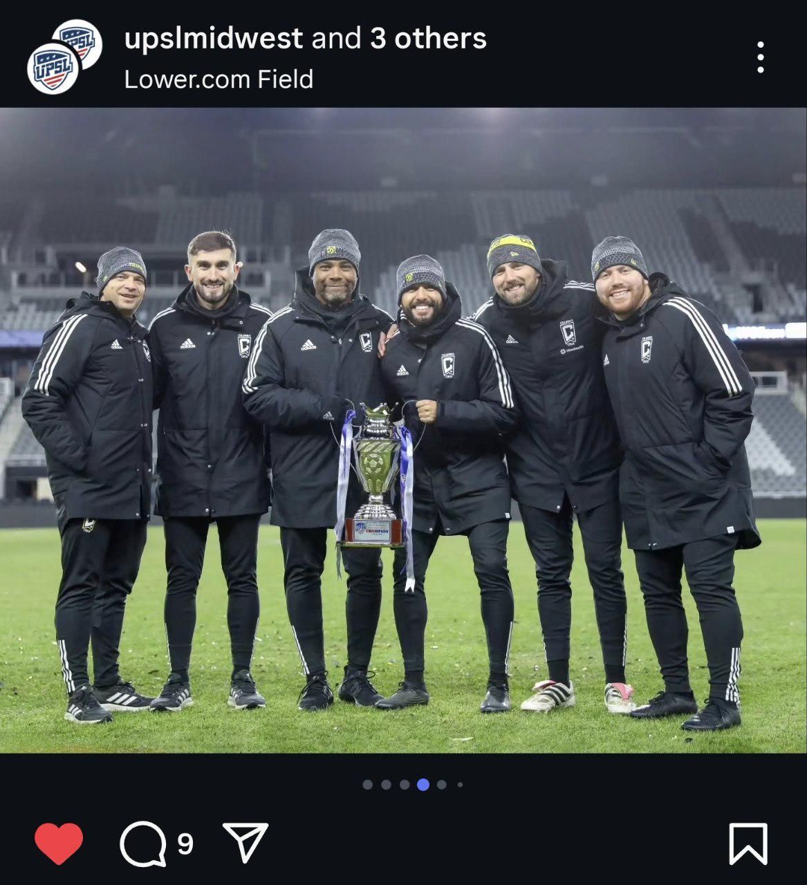Group of coaches posing with trophy on field at Lower.com Field at night.