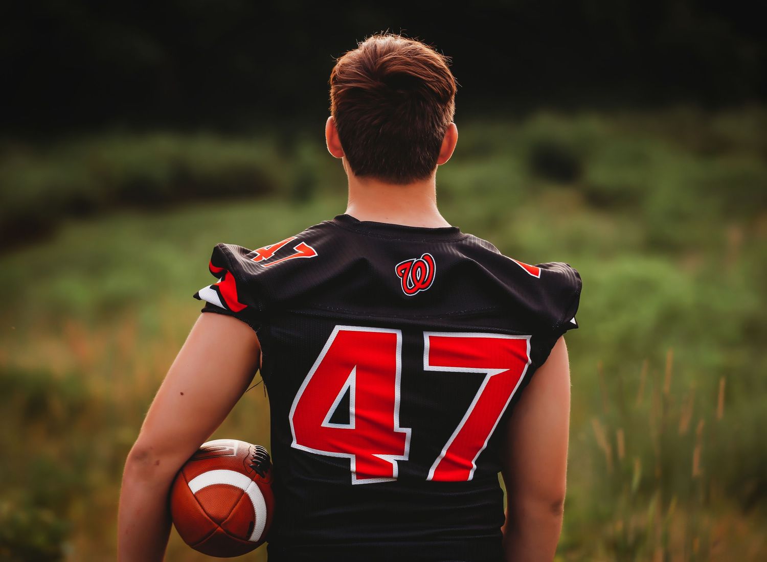 Football player wearing black jersey with number 47 stands in grassy field at sunset.