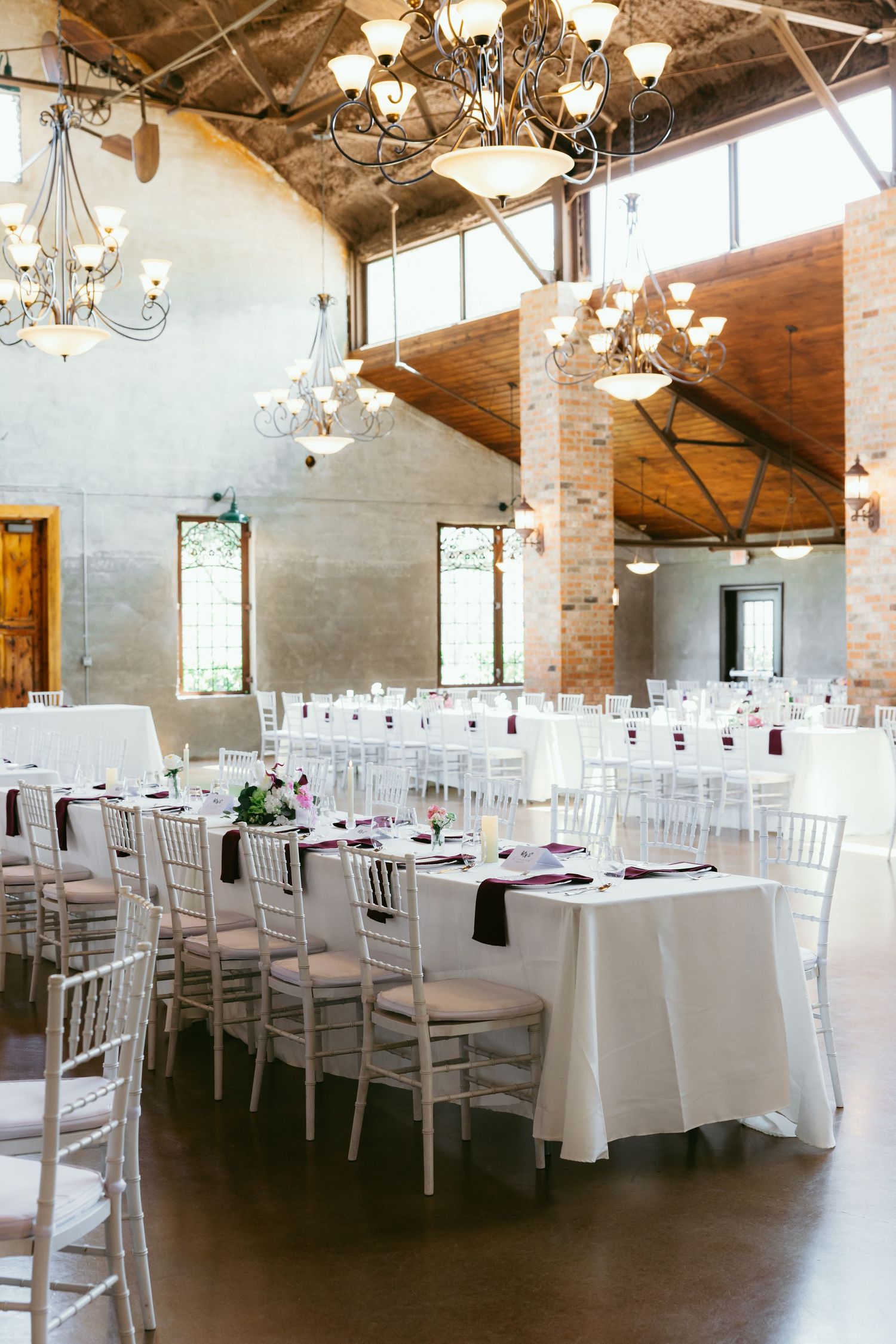 Elegant wedding reception hall with white Chiavari chairs, long tables, and rustic brick columns under chandelier lighting.