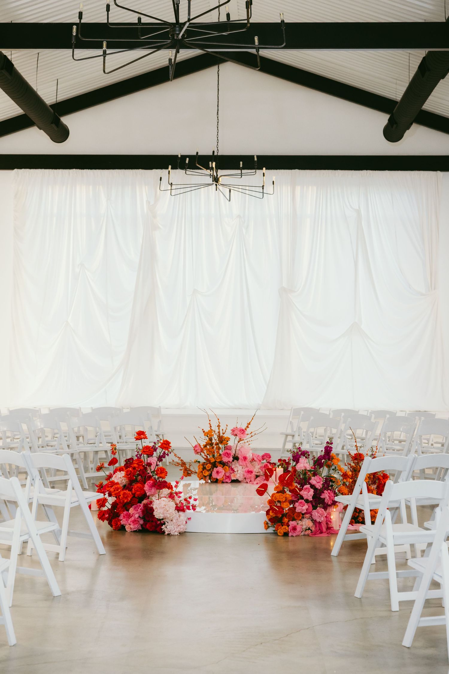Elegant wedding ceremony setup with white chairs, vibrant pink and red floral arrangements, and draped white backdrop.