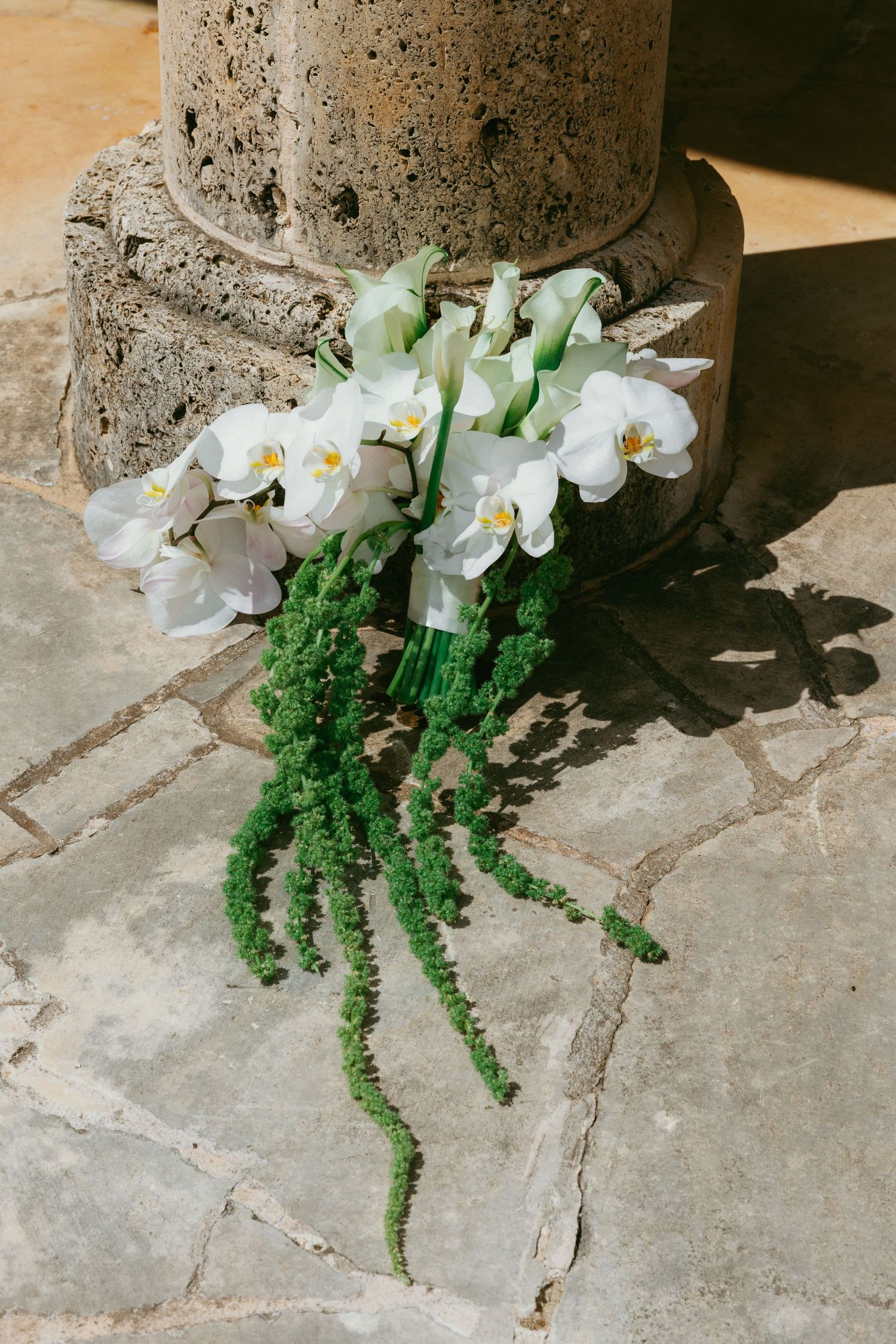White orchid and calla lily bouquet with green trailing amaranthus resting against a stone column on flagstone.
