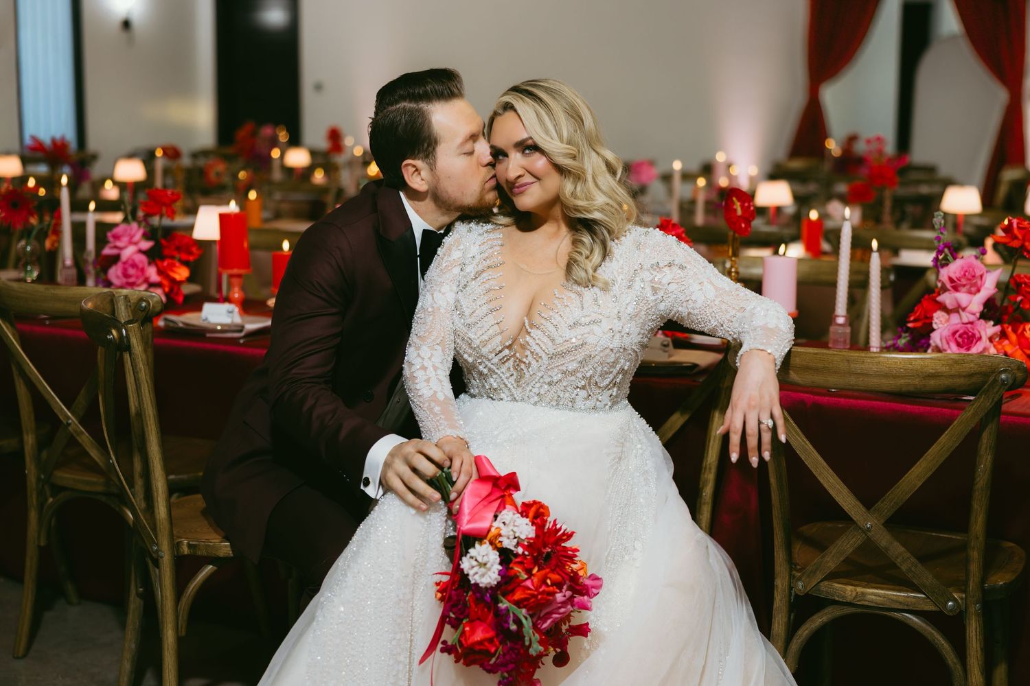 A wedding celebration with red and pink floral decor featuring elegant table settings and rustic wooden chairs.