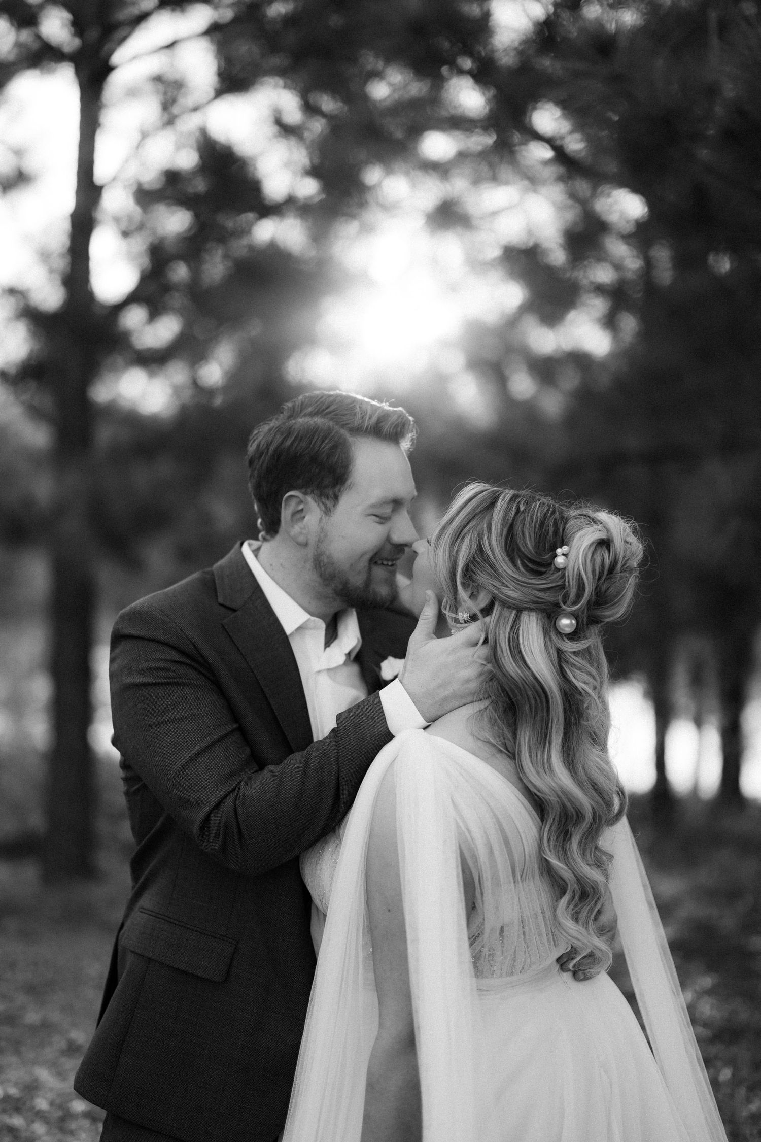 A romantic black and white wedding portrait shows a couple sharing a tender kiss in a wooded outdoor setting.
