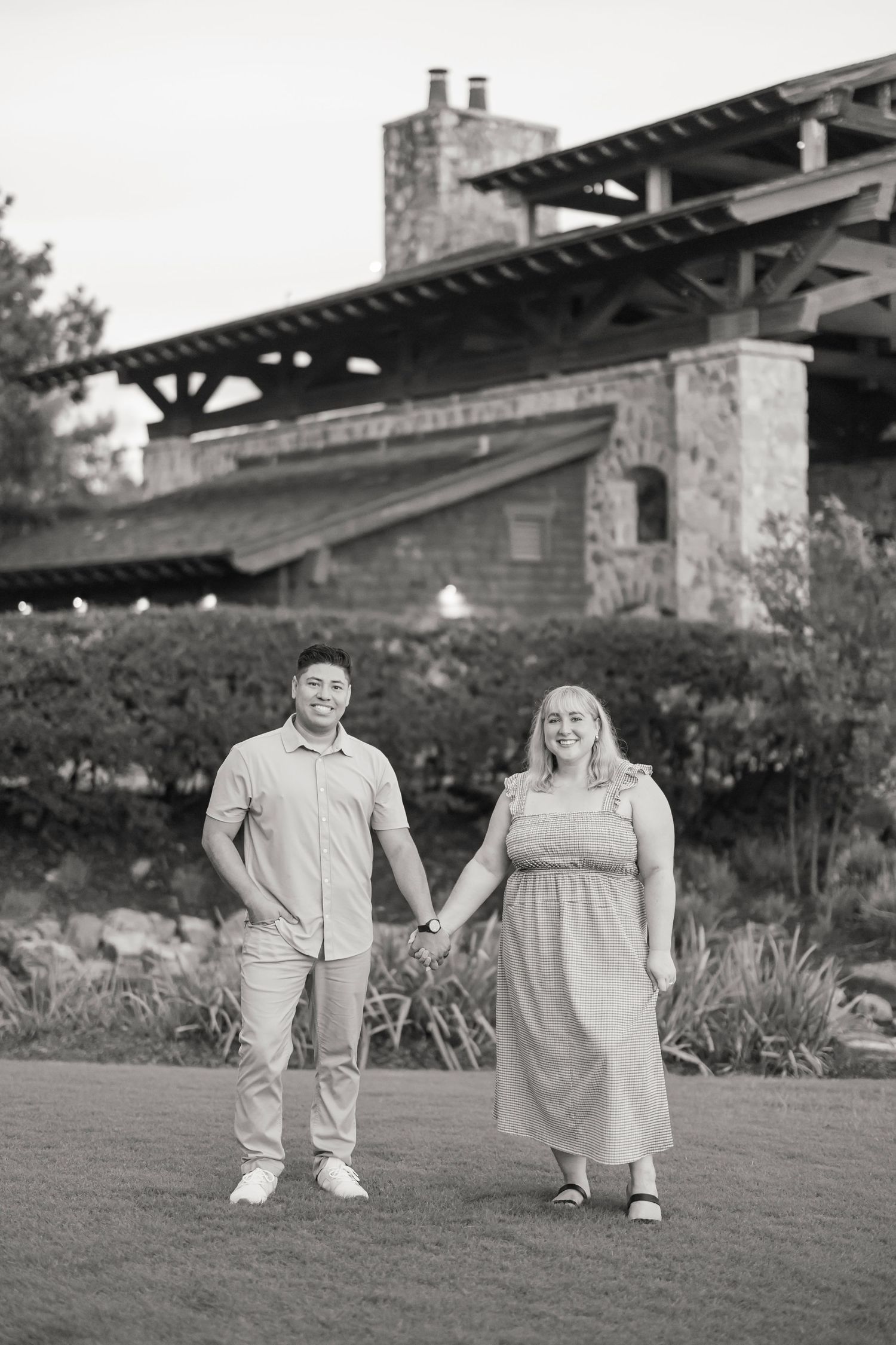 Couple holding hands smiling in front of rustic stone building with lush greenery in black and white photo.