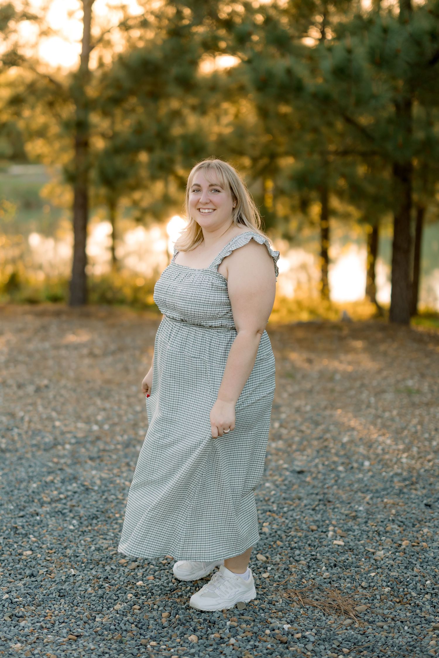 A person in a long gingham dress smiles while standing on gravel during sunset.