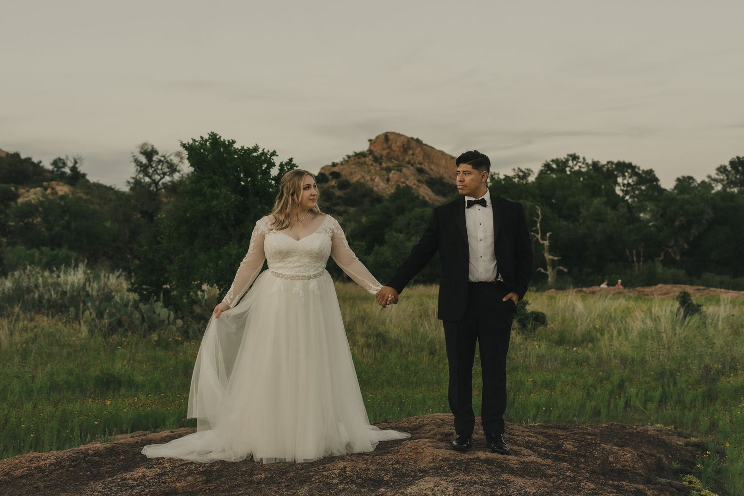 Bride in white gown and groom in tuxedo hold hands on rocky landscape with green meadow and hills at dusk.