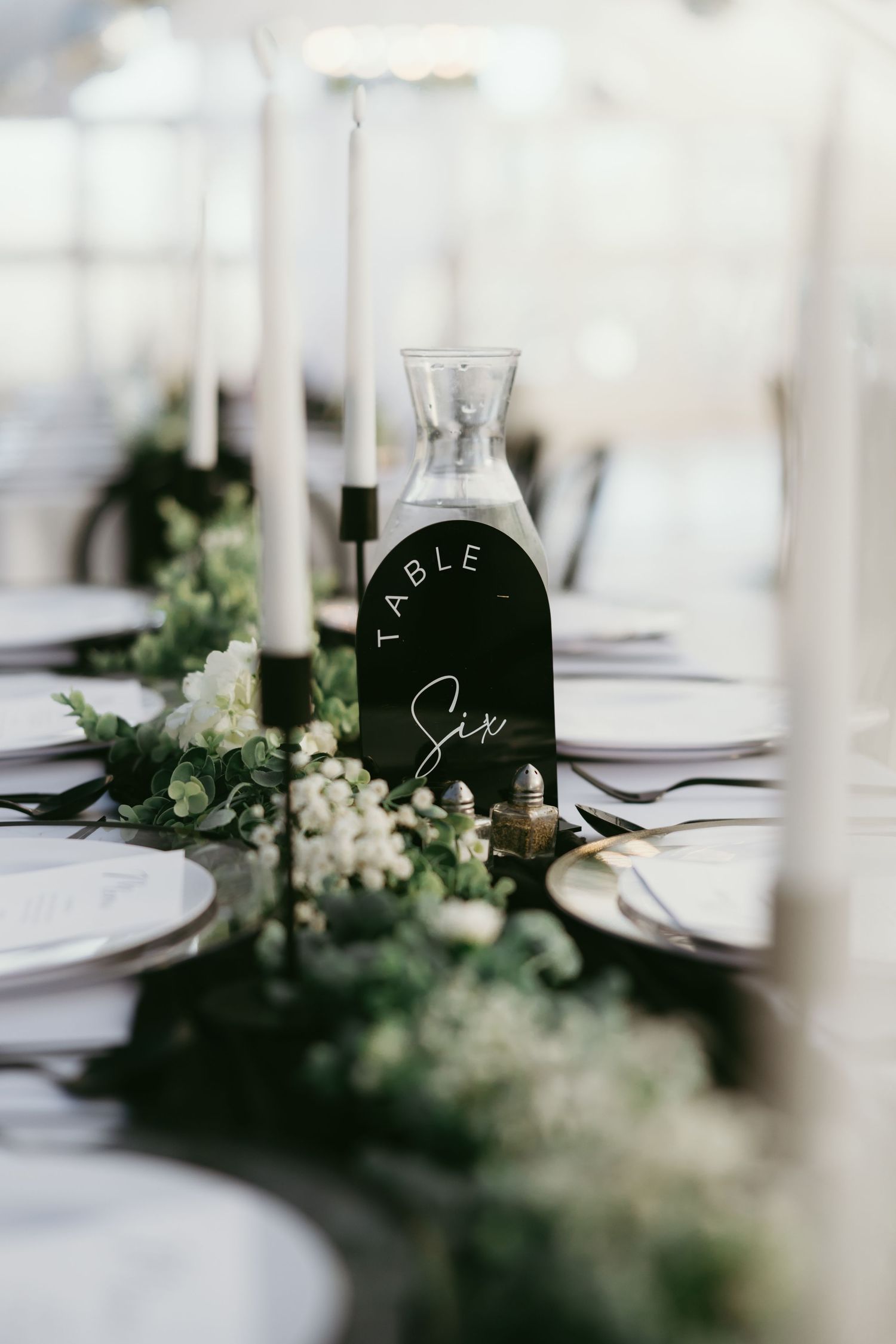 Elegant wedding table setting with white candles, greenery garland, and a black arch Table Six sign centerpiece.
