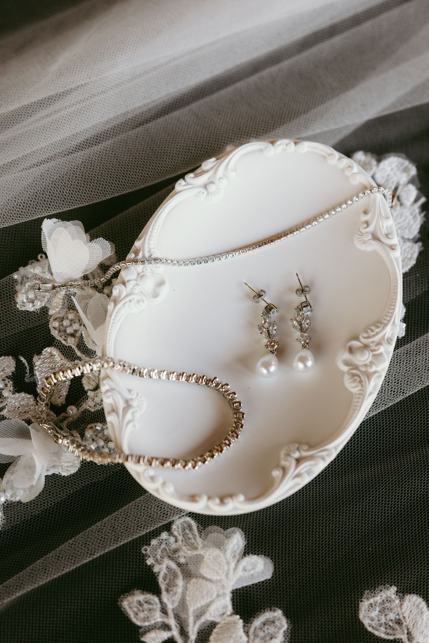 Elegant bridal jewelry set with pearl drop earrings and crystal necklace displayed on white ornate dish with lace veil.