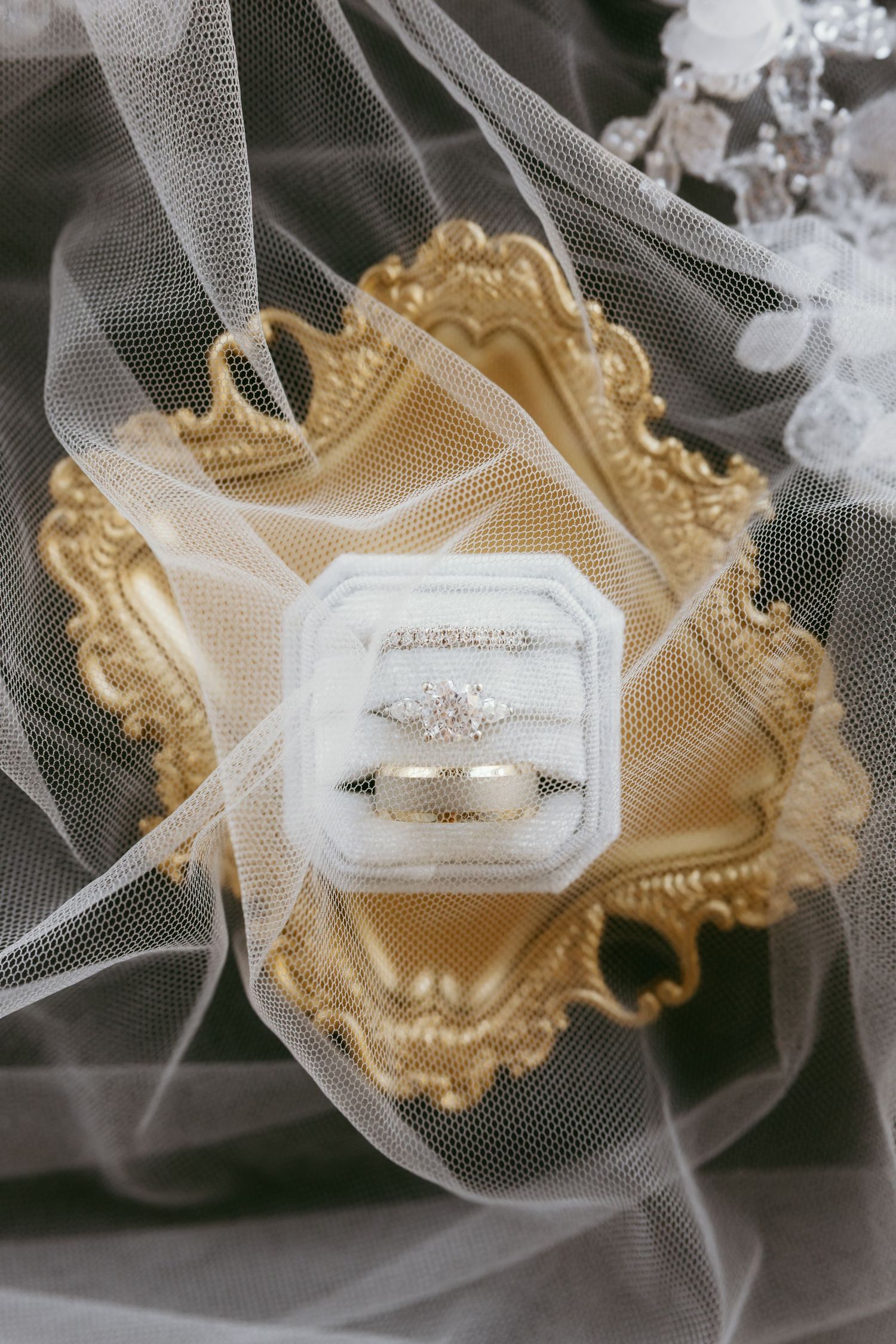 Wedding rings displayed in white velvet box on ornate gold tray with bridal veil, featuring diamond engagement ring and bands.