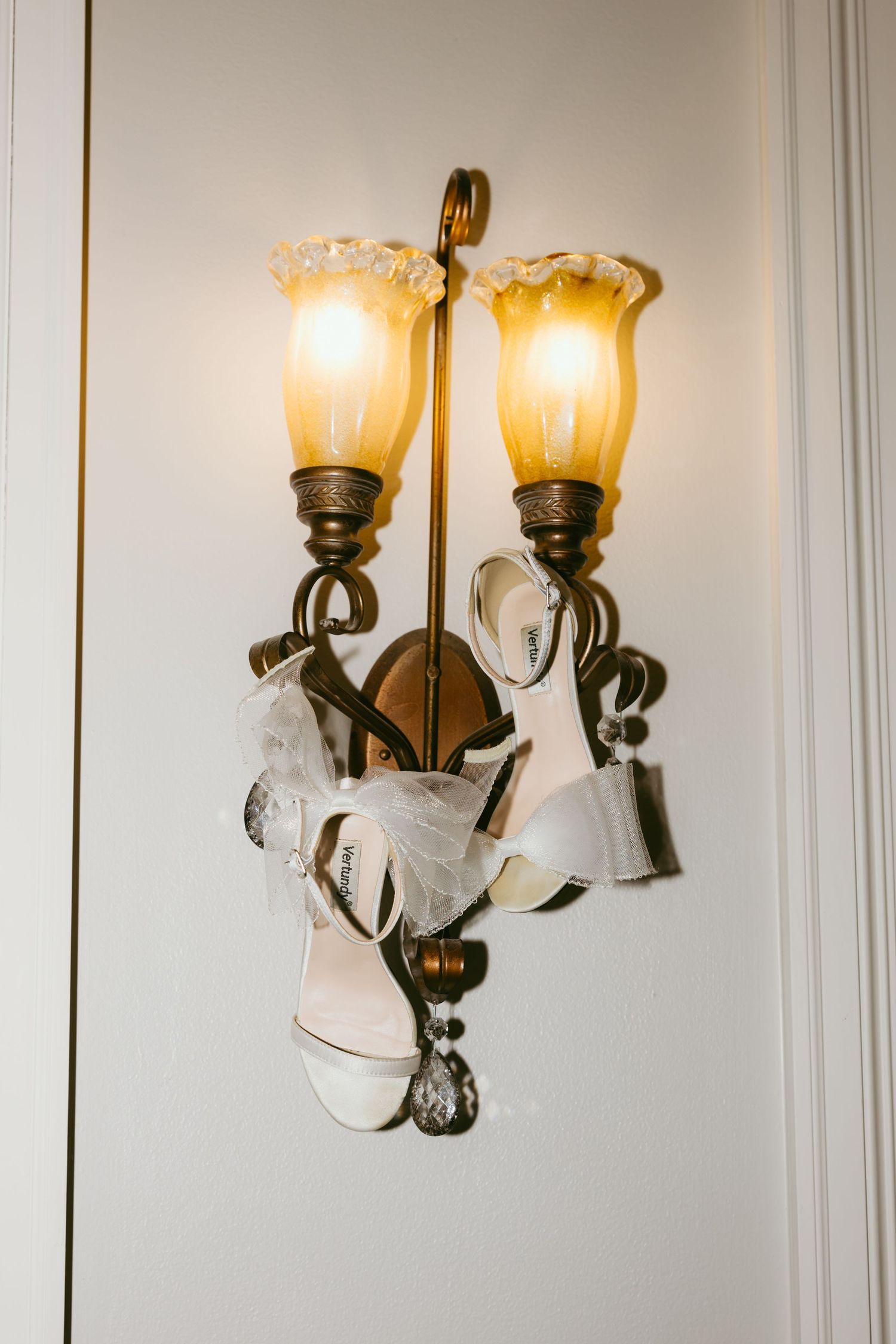 White bridal heels with tulle bow hanging on ornate brass wall sconce with amber glass shades.