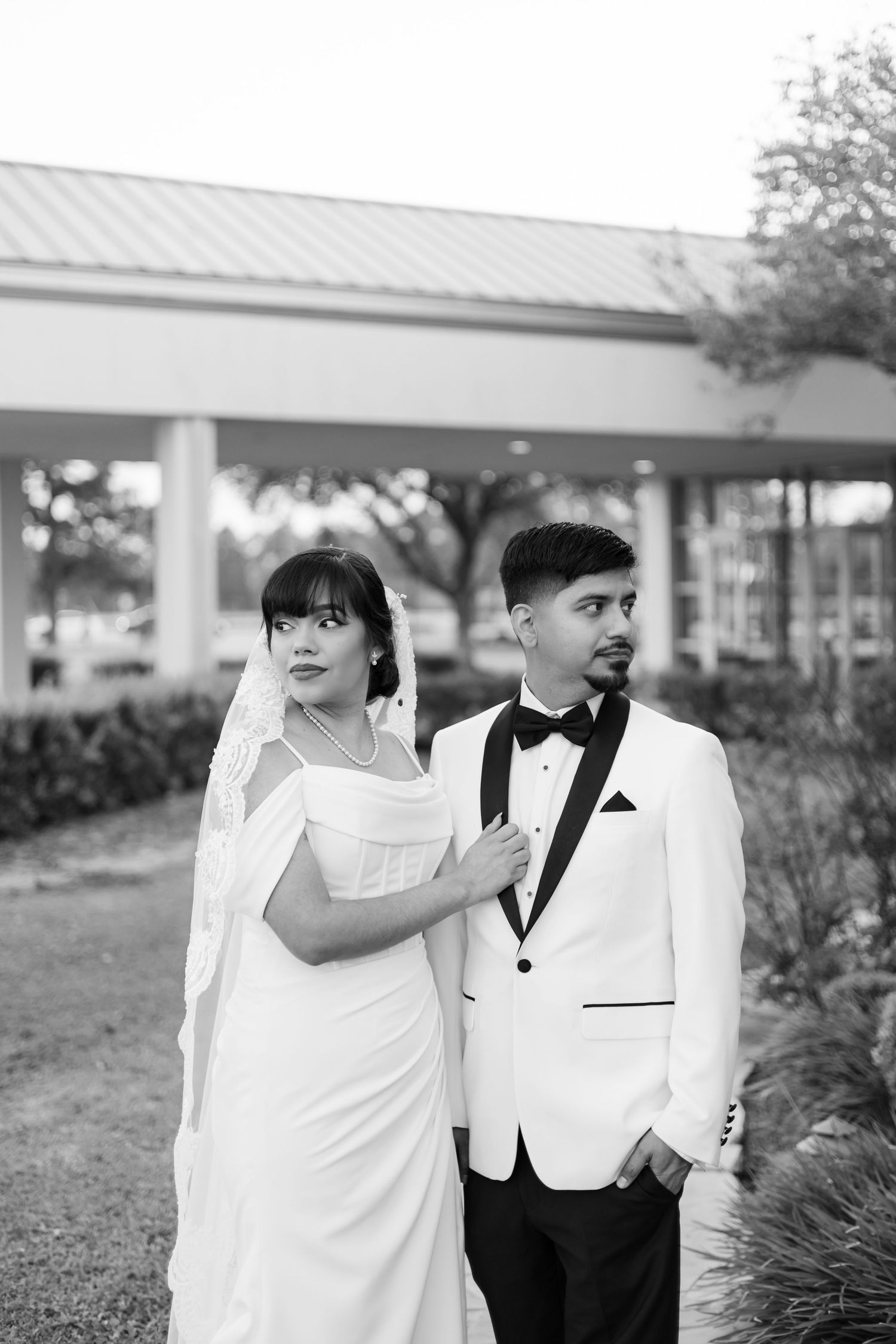 Black and white photo of a bride in white gown and groom in tuxedo posing elegantly outdoors at their wedding.