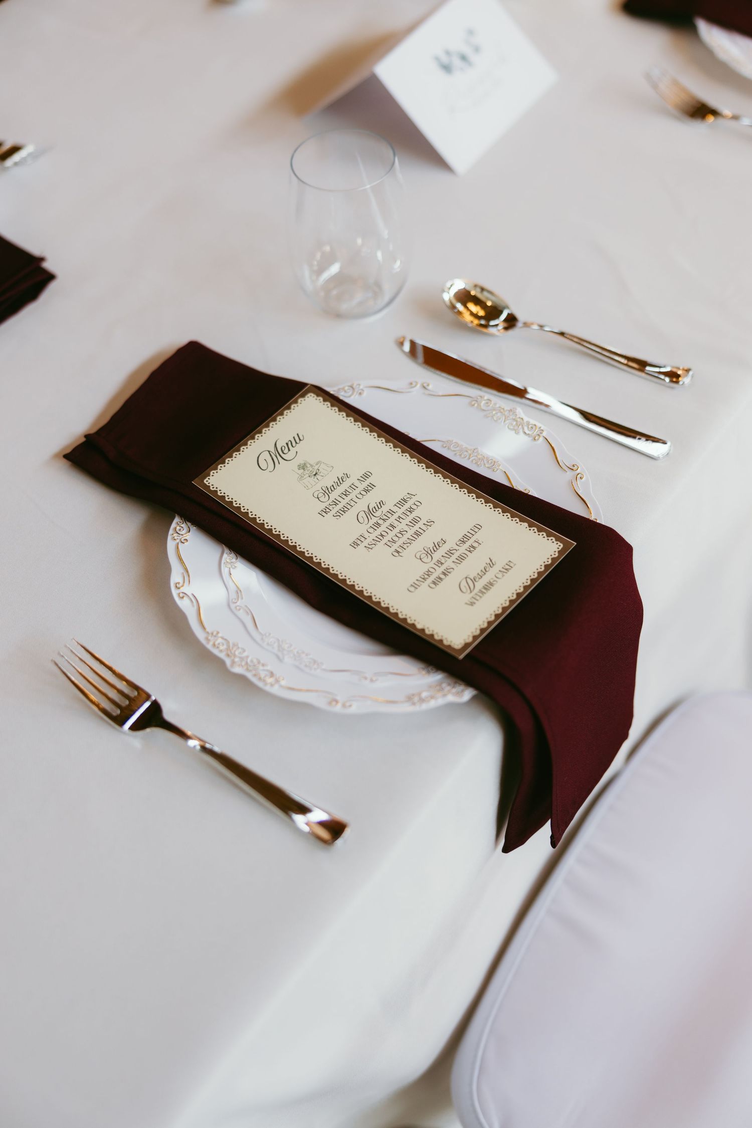 Formal wedding place setting with burgundy napkin, gold cutlery, ornate white plate, and elegant menu card on white tablecloth.