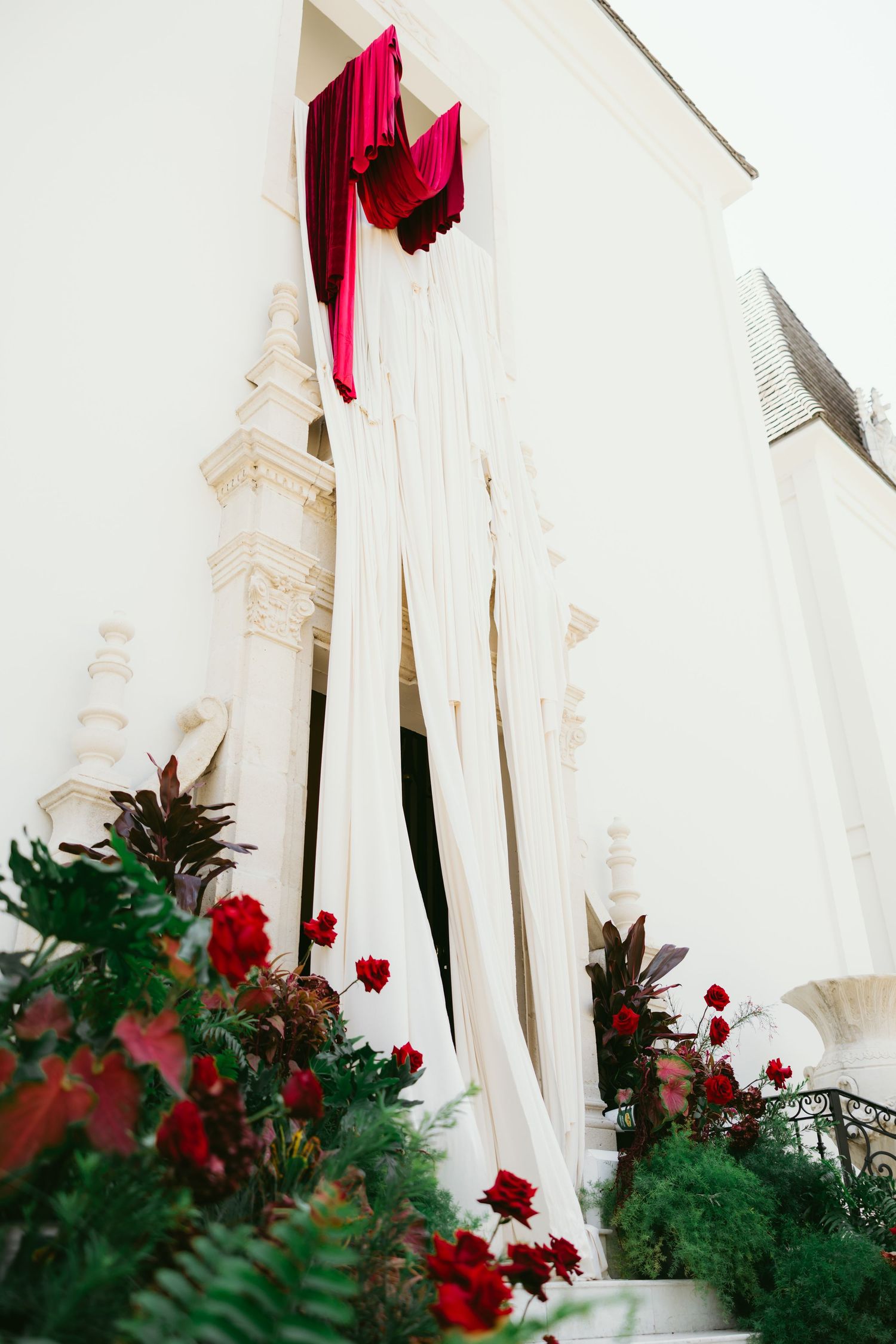 White draped fabric with red velvet accents adorns a classical building entrance, flanked by lush greenery and red roses.