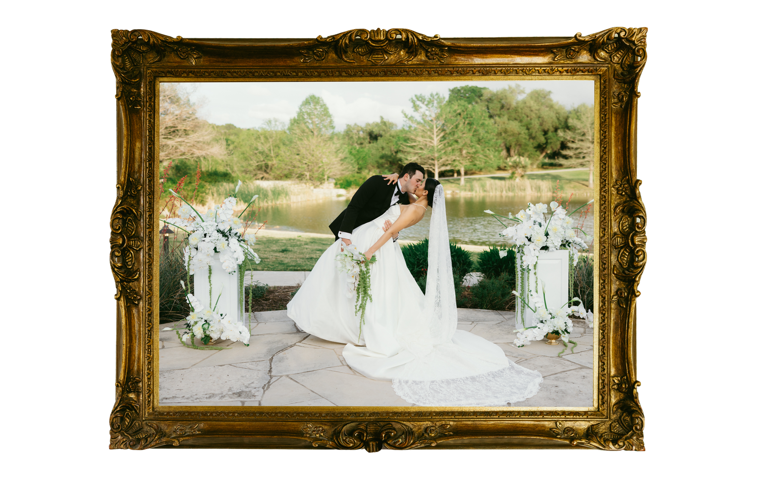 Bride and groom kissing at outdoor wedding ceremony by a lake, framed in ornate gold frame with white floral arrangements.