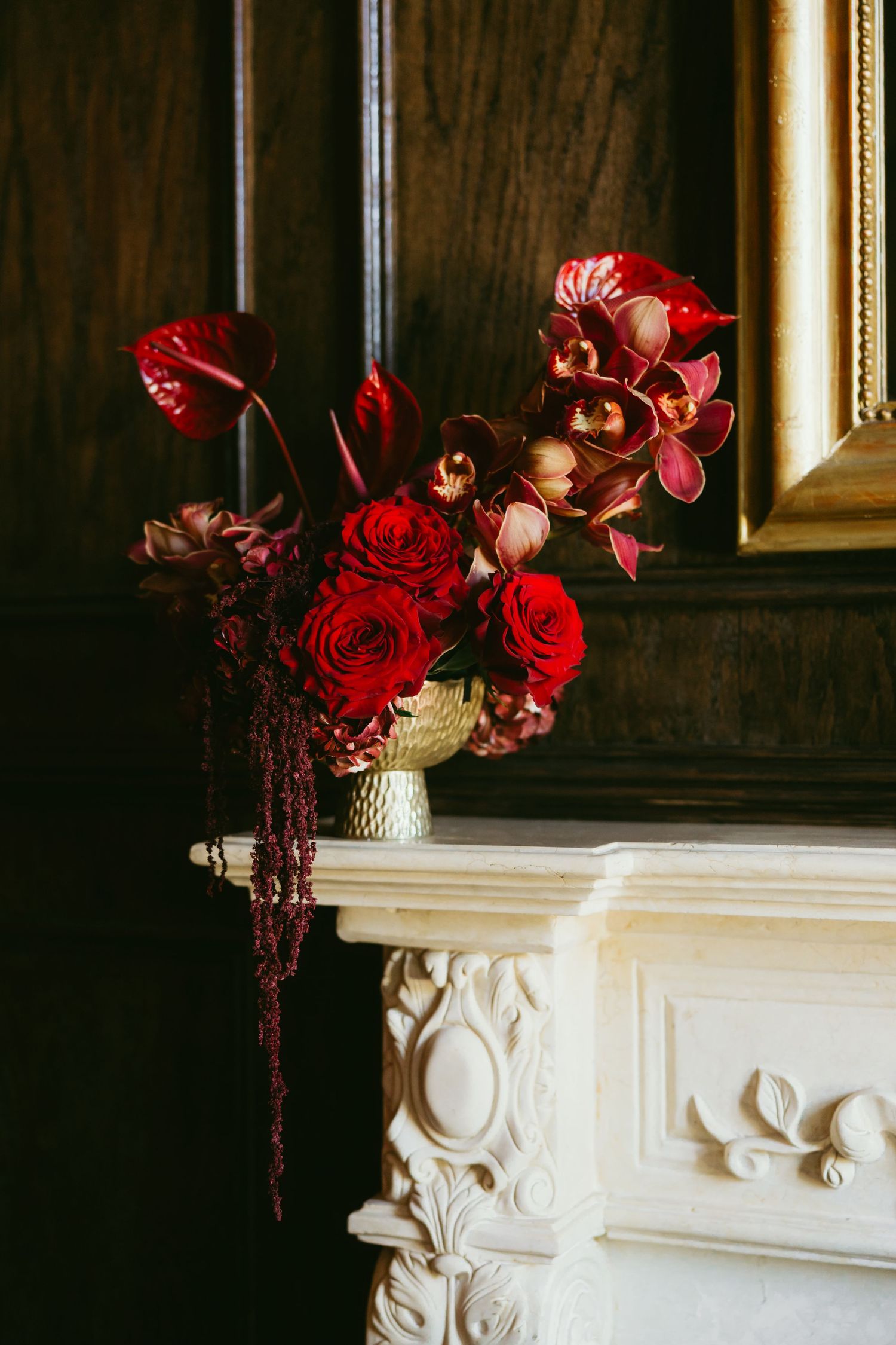 Red roses and orchids in a gold vase on a white ornate fireplace mantel against dark wood paneling.