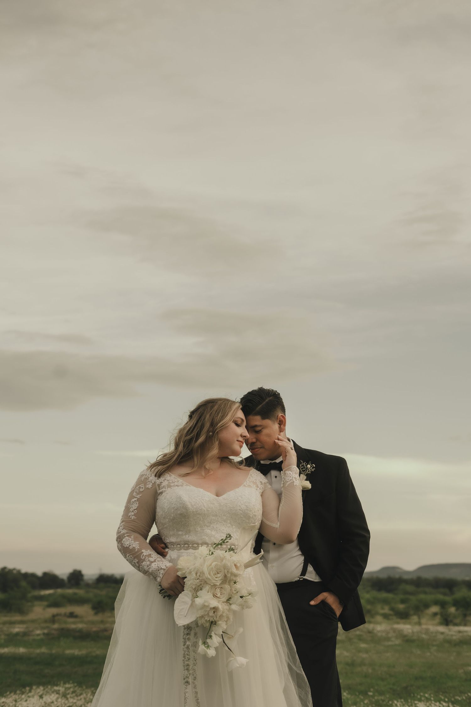 Bride in lace gown holding white bouquet stands with tuxedo-clad groom in scenic outdoor wedding portrait.