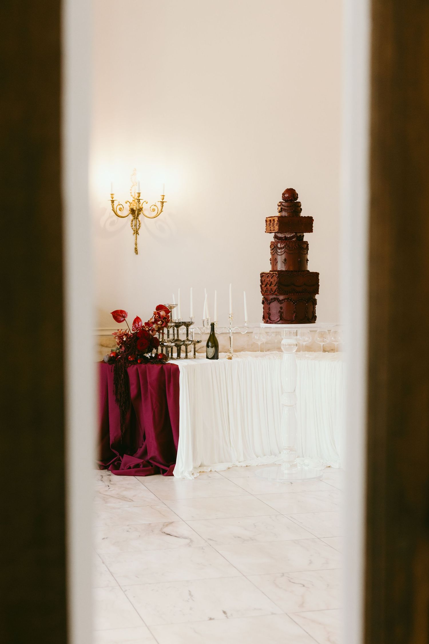 Elegant wedding reception with a tall chocolate cake, burgundy draped table, red florals, candles, and a gold wall sconce.