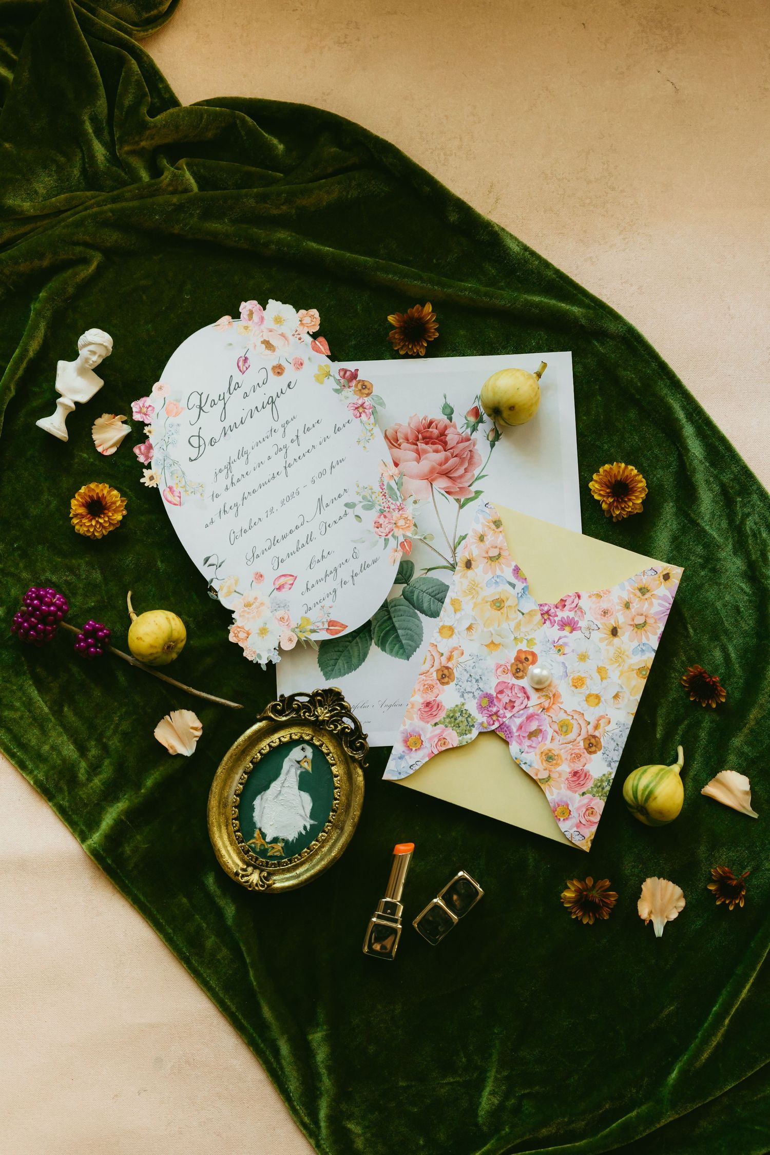 Floral wedding invitation suite on green velvet with vintage frame, lipstick, dried flowers, and small fruits.