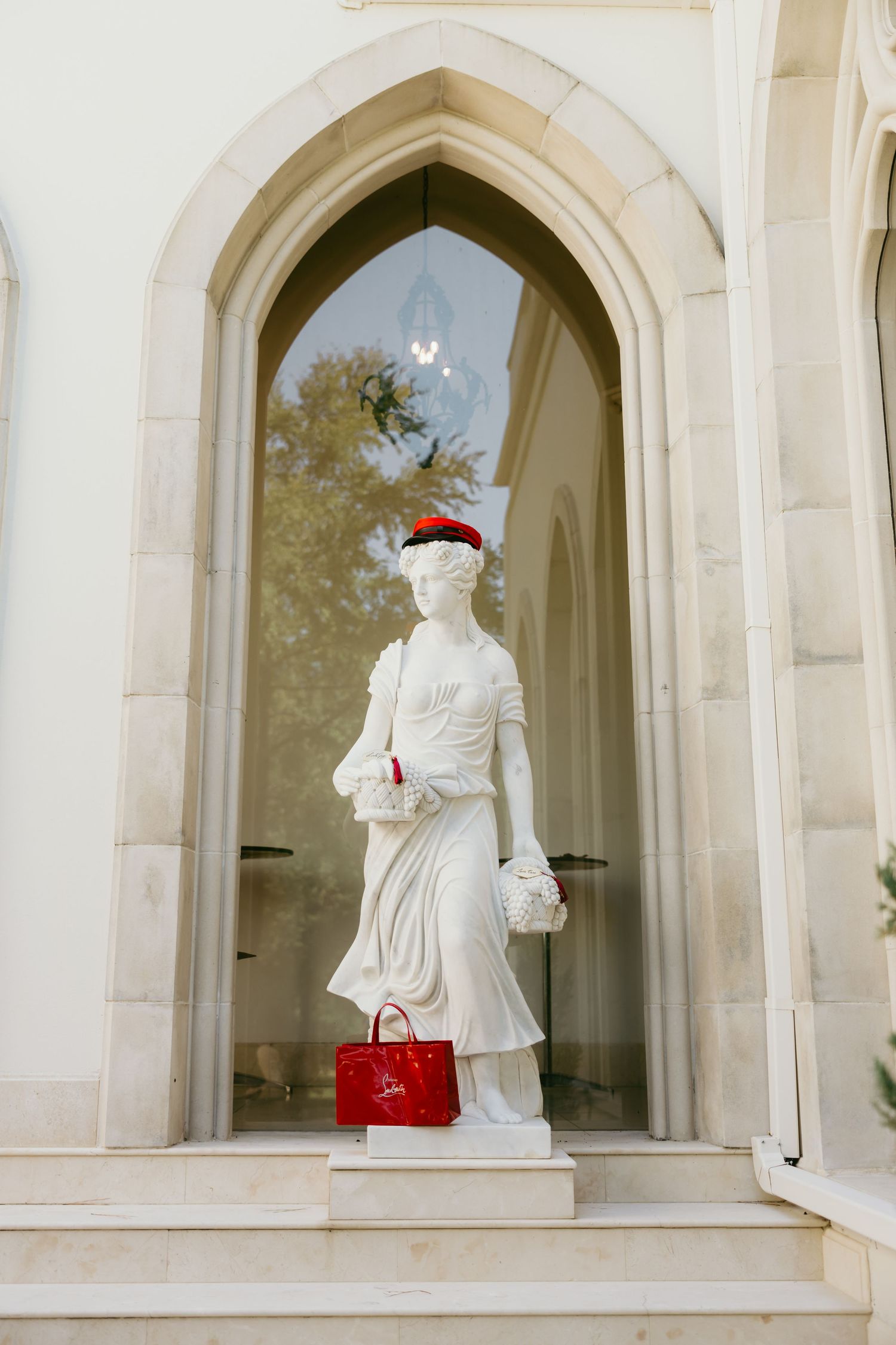 White classical statue adorned with red handbag and floral hat, displayed in Gothic arch alcove of elegant building.