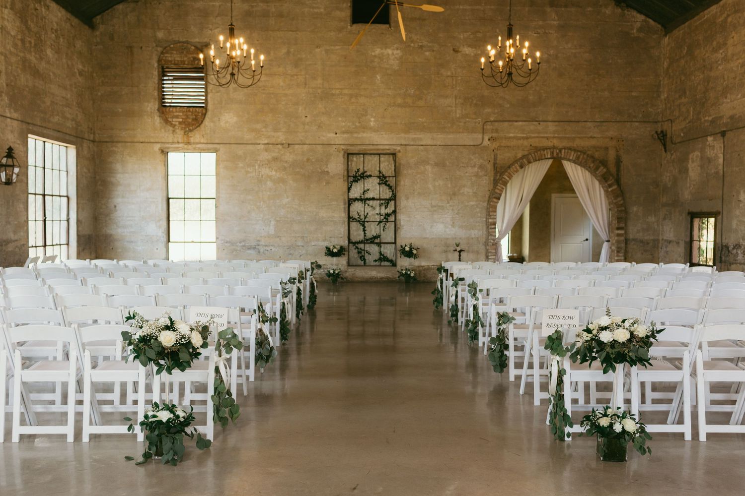 Elegant wedding ceremony setup in a rustic stone venue with white chairs, floral arrangements, and chandeliers.