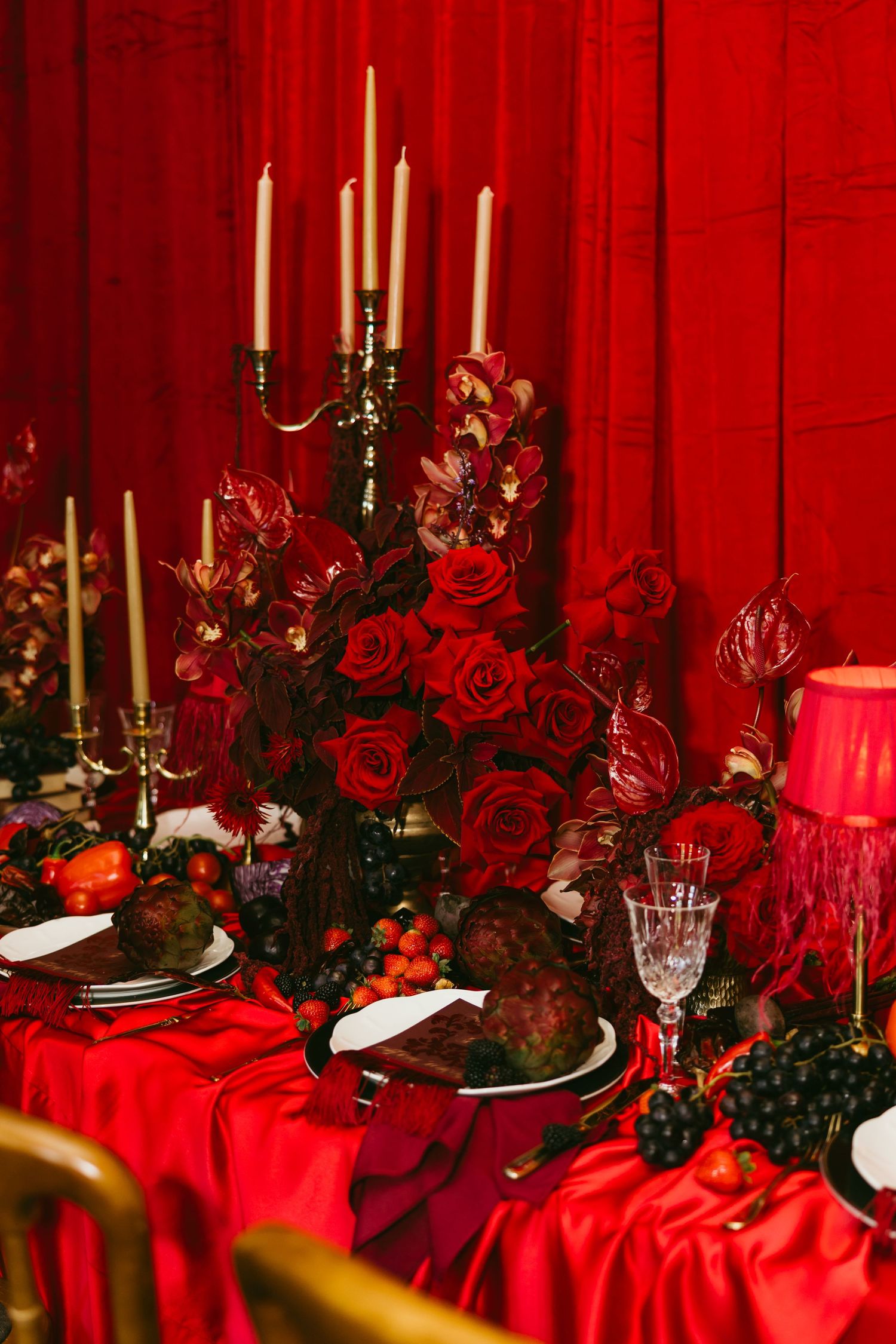 Elegant red tablescape with roses, candelabra, crystal glasses, and dark fruits on a crimson satin tablecloth.