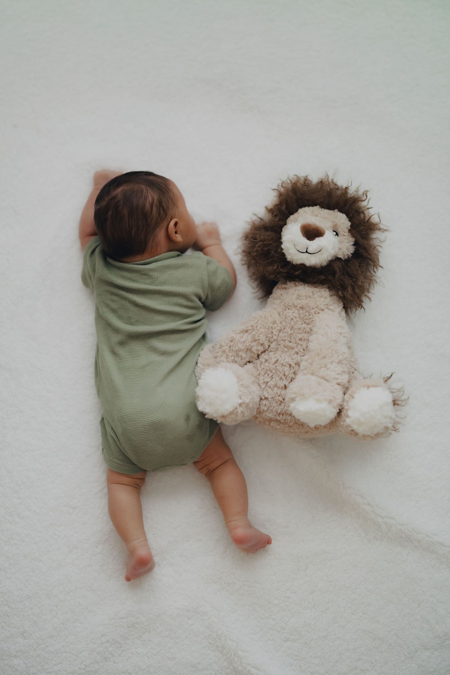 Newborn baby in green onesie sleeping beside a lion stuffed animal on a white blanket.