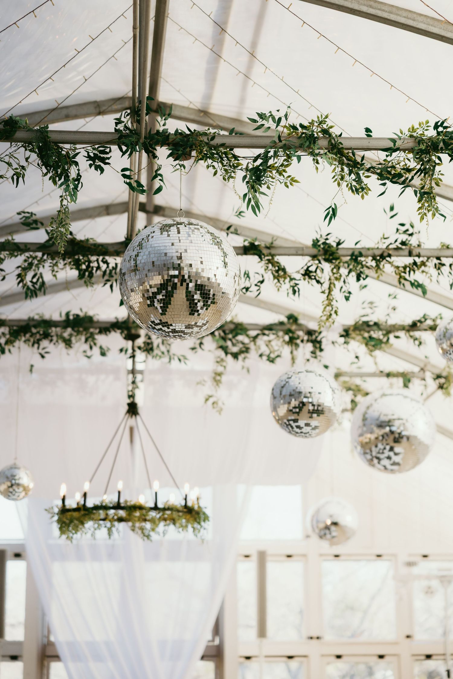 Elegant wedding tent ceiling with disco balls, greenery garlands, and a floral chandelier with candles.
