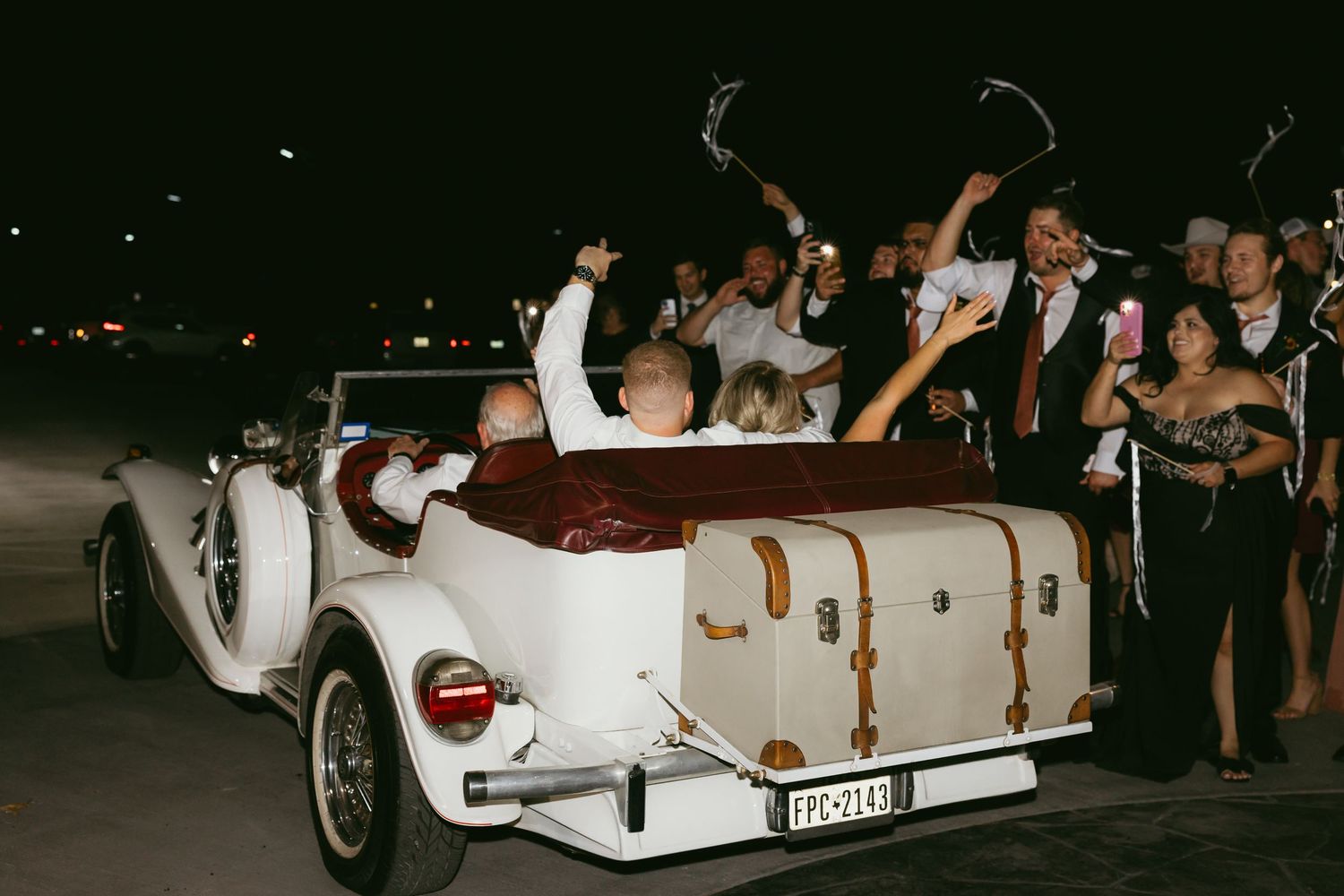 Wedding guests celebrate around a vintage white convertible car during a nighttime celebration.