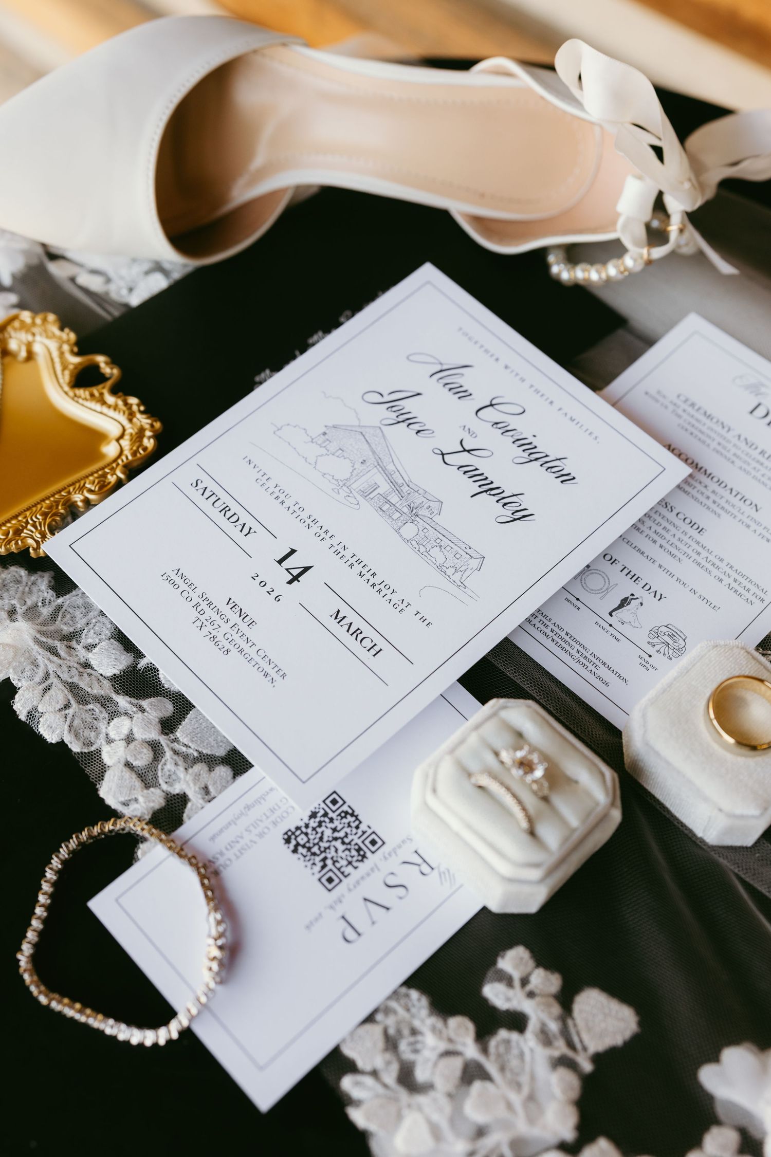 Elegant wedding flat lay with white heels, pearl bracelet, invitation suite, ring box, and gold tray on black lace fabric.