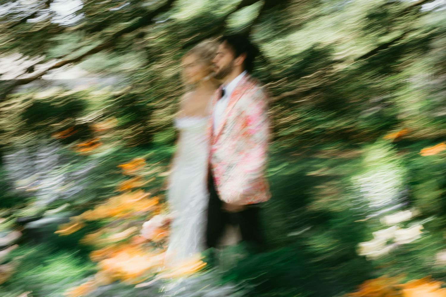 Blurred artistic wedding photo of bride in white dress and groom in floral suit embracing among lush green foliage.