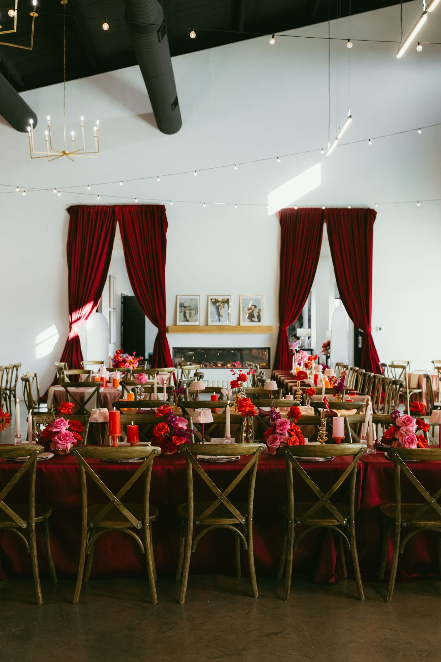 Elegant wedding reception with red velvet drapes, long tables adorned with red and pink floral centerpieces, and warm string lights.