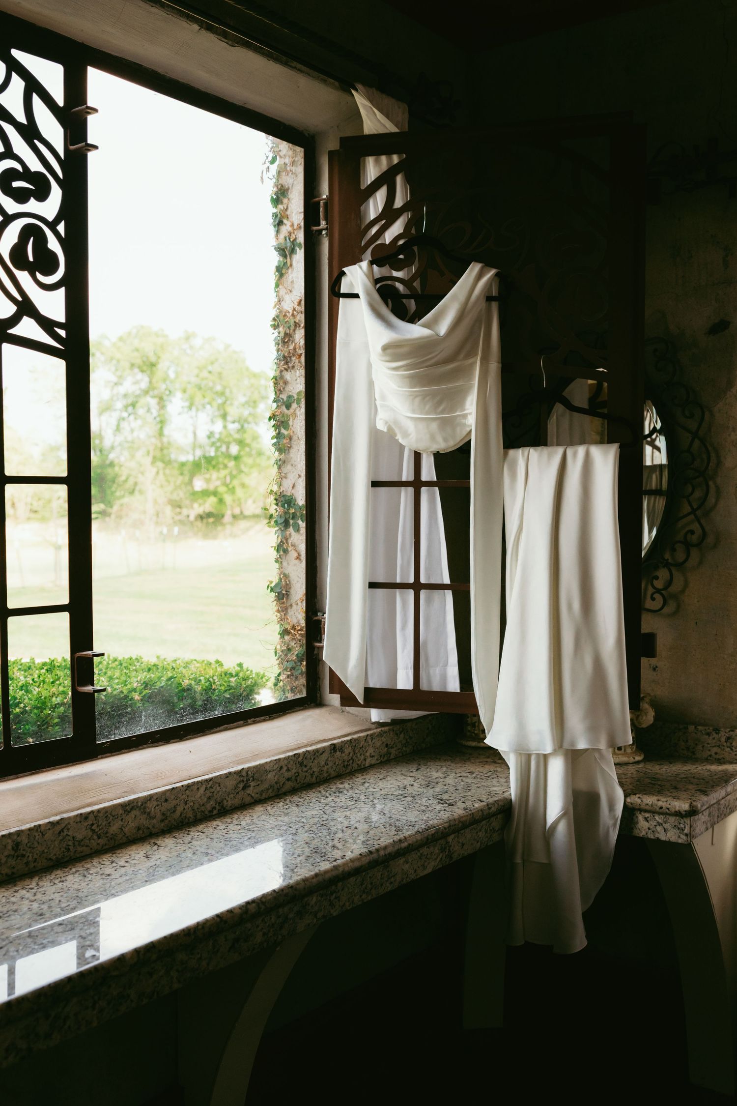 White wedding dress hanging near ornate window with garden view in elegant rustic venue.