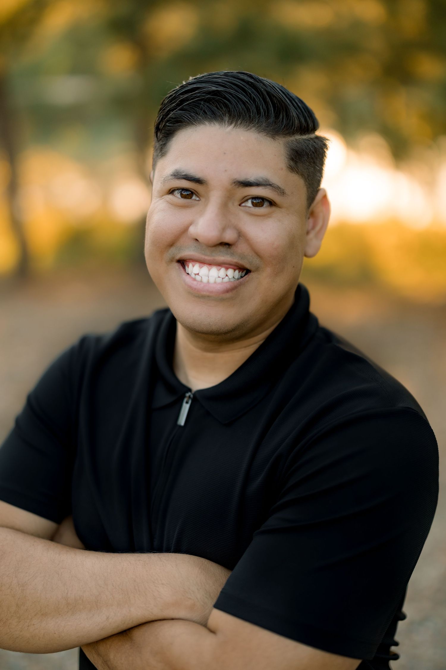 A person wearing a black polo smiles warmly in an outdoor portrait setting.