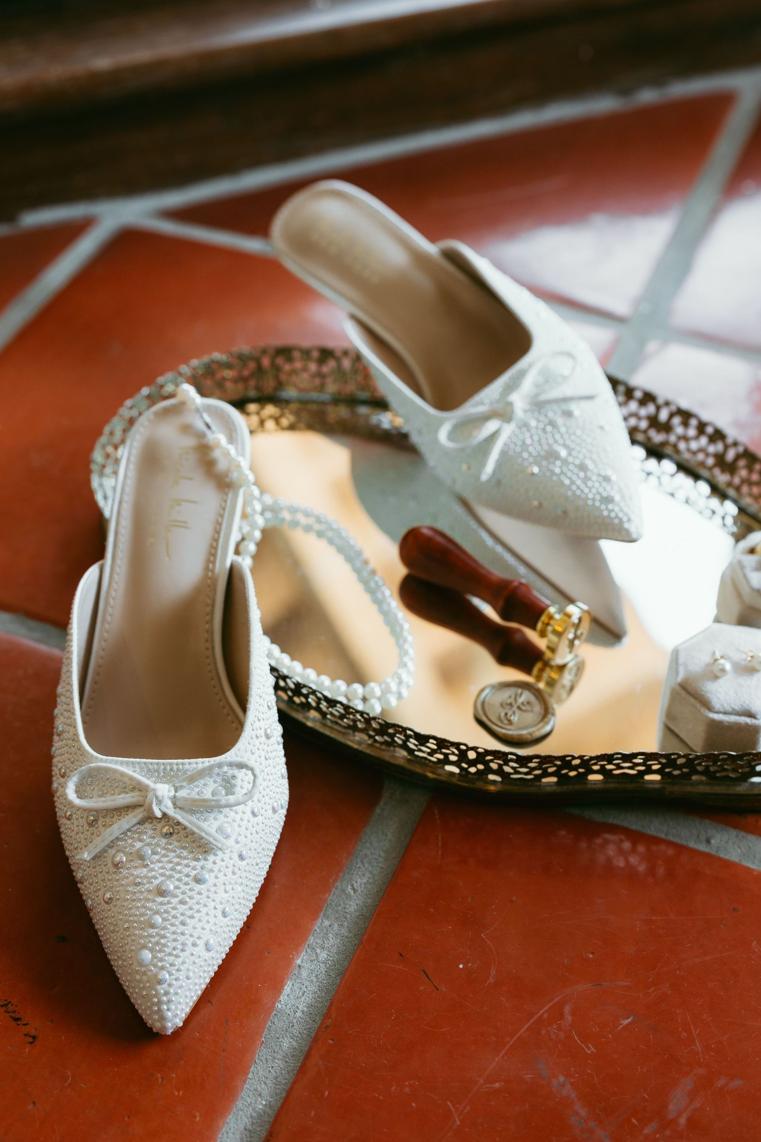 Pearl-embellished white bridal slingback heels displayed on a decorative tray with rings and accessories on terracotta tiles.