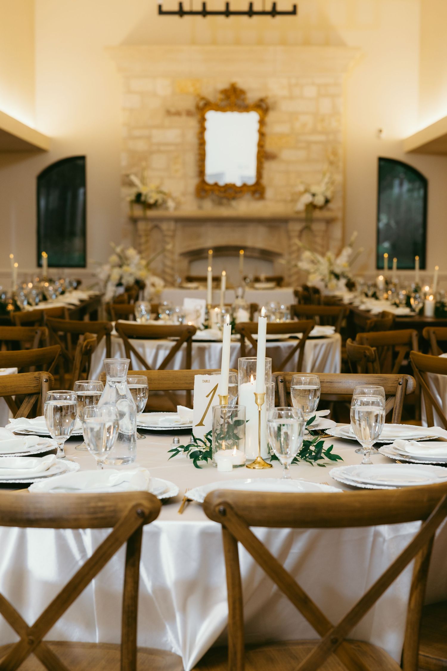 Elegant wedding reception hall with white tablecloths, wood cross-back chairs, candles, and a stone fireplace backdrop.
