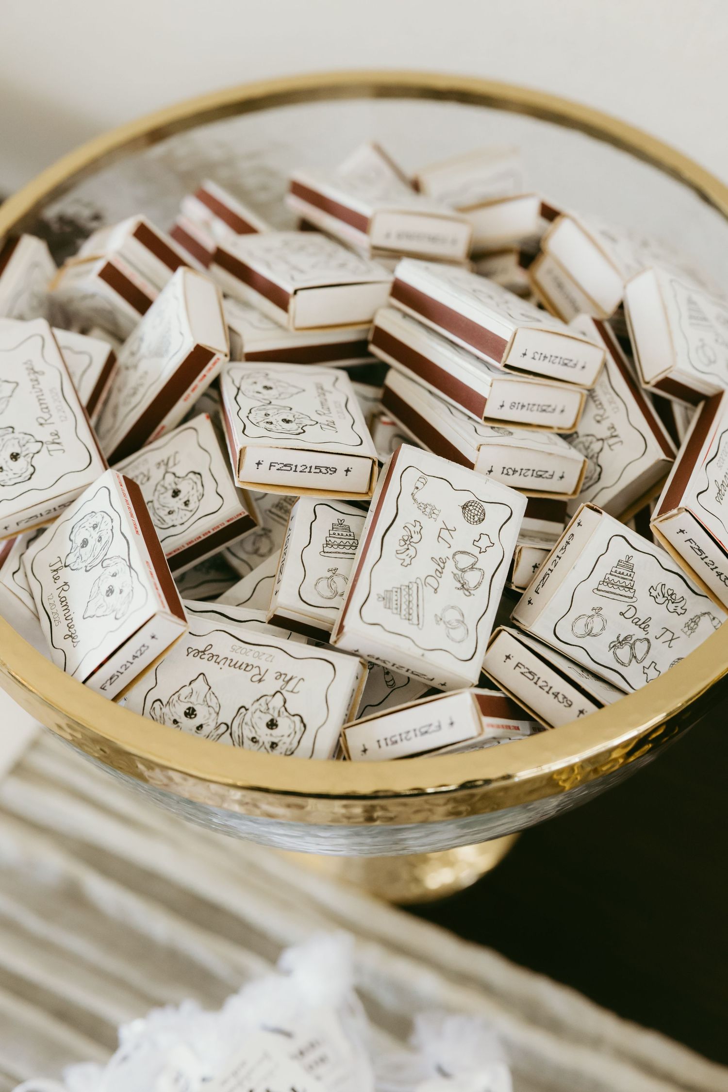 Custom illustrated matchboxes with wedding designs displayed in a gold pedestal bowl as elegant party favors.