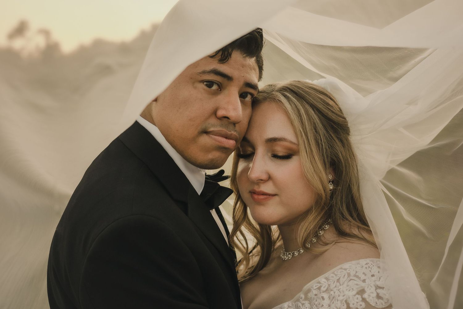 Bride and groom share an intimate moment under a flowing veil at golden hour in a romantic wedding portrait.