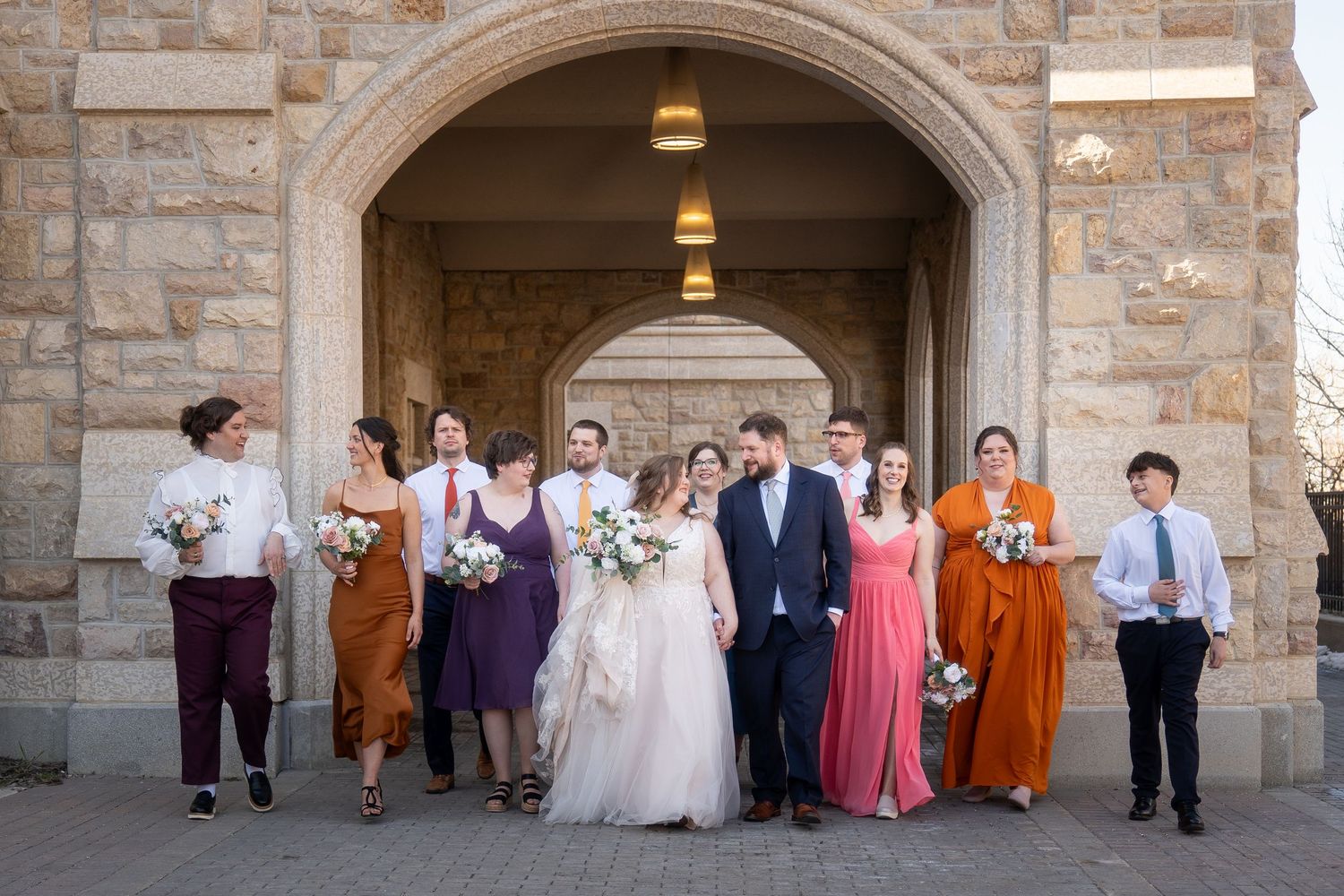 Wedding party members stand under stone archways in coordinated formal attire during outdoor photo session.