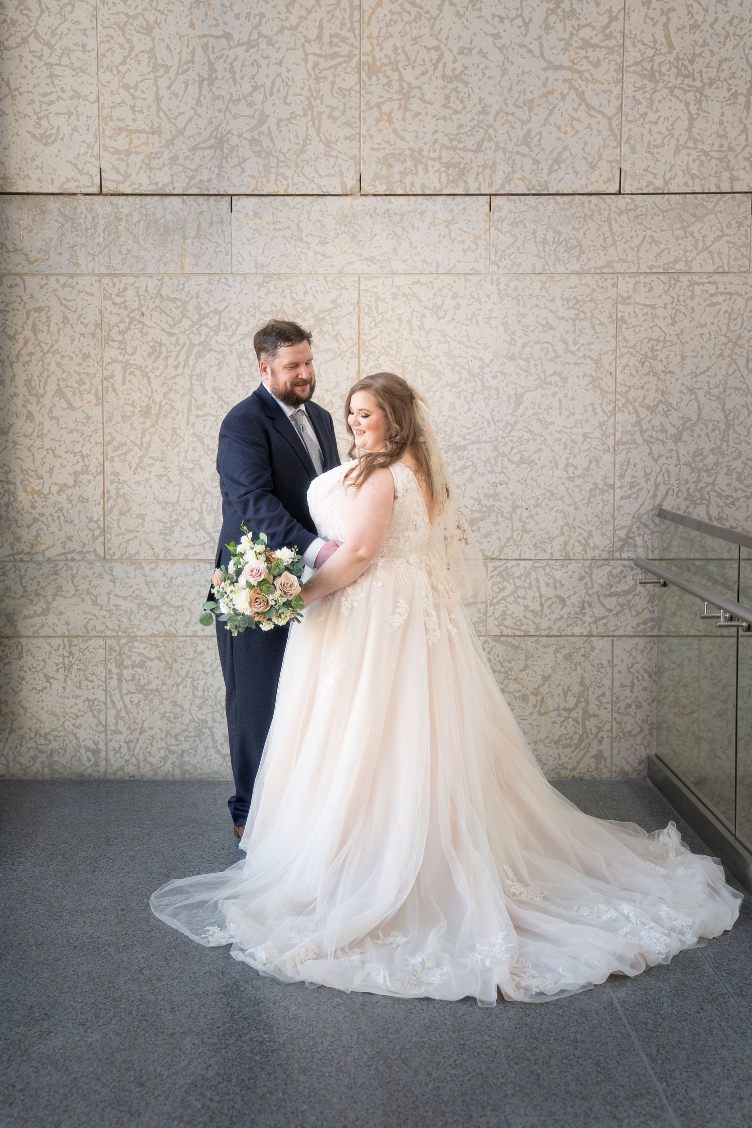 A bride in a flowing white ballgown and groom in navy suit share an intimate moment against a textured wall.