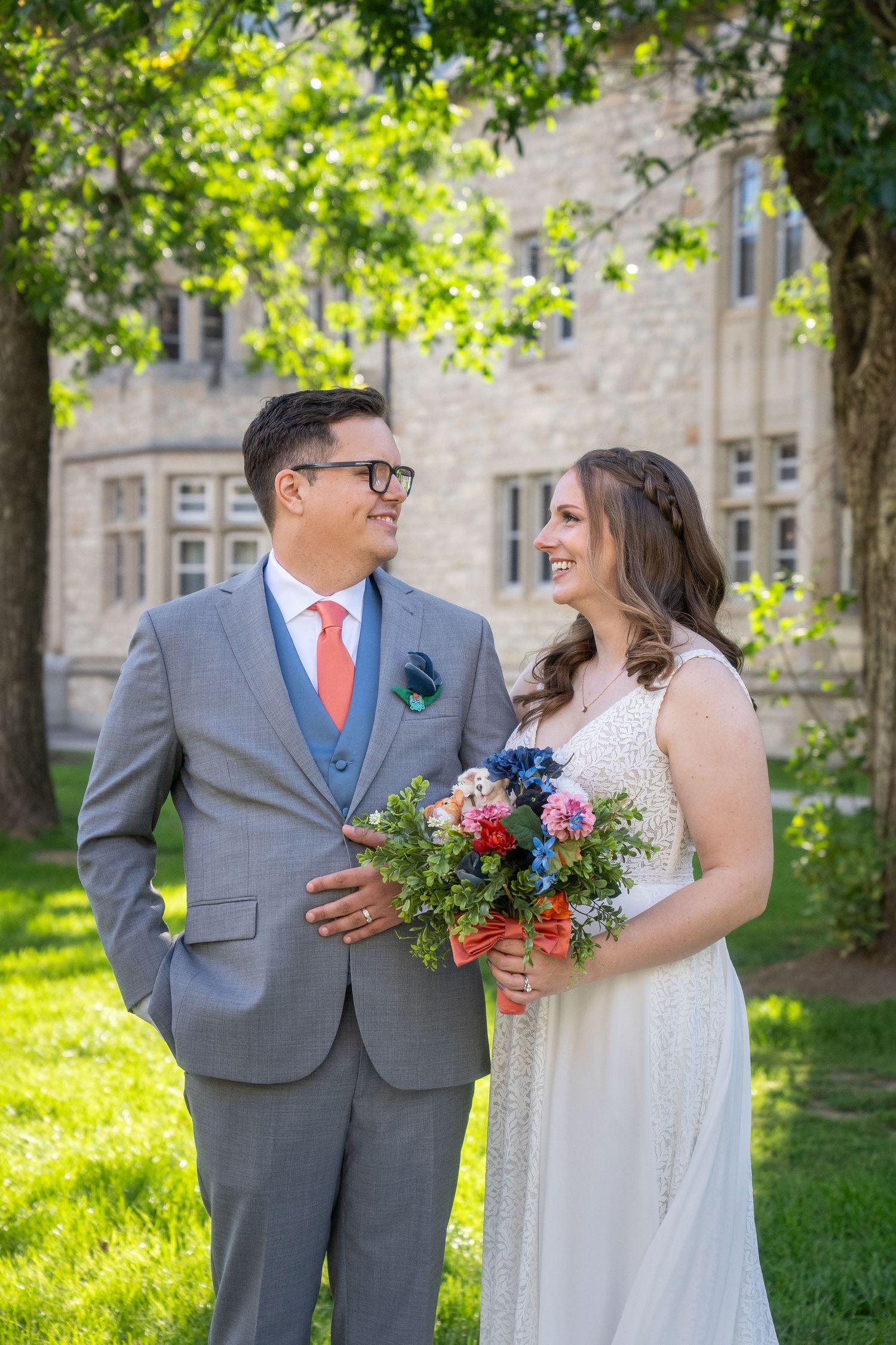 A couple in wedding attire stand together on a sunny day, with the bride holding a colorful bouquet against stone building.