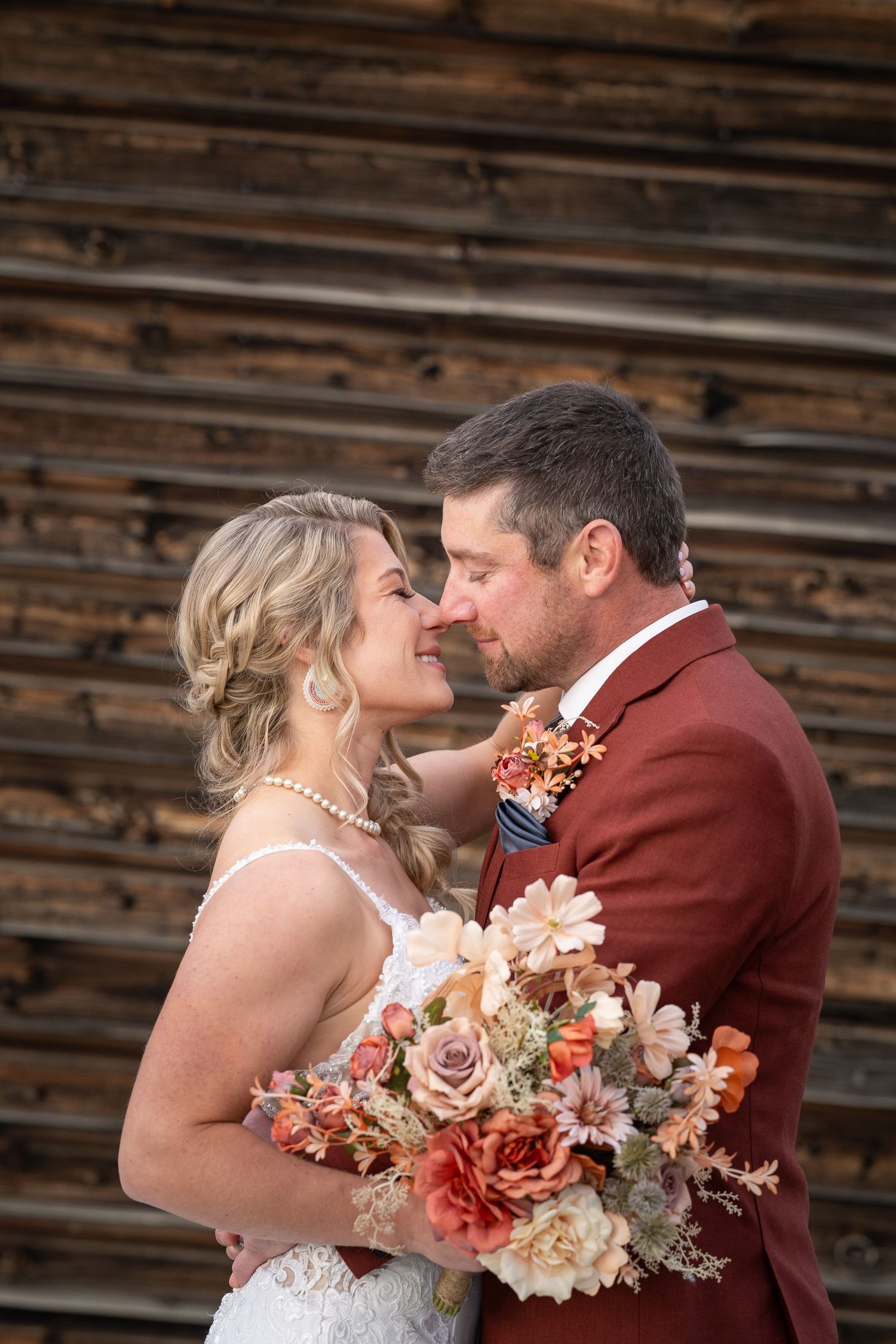 Romantic wedding couple shares intimate moment against rustic wooden wall with autumn-colored floral bouquet.