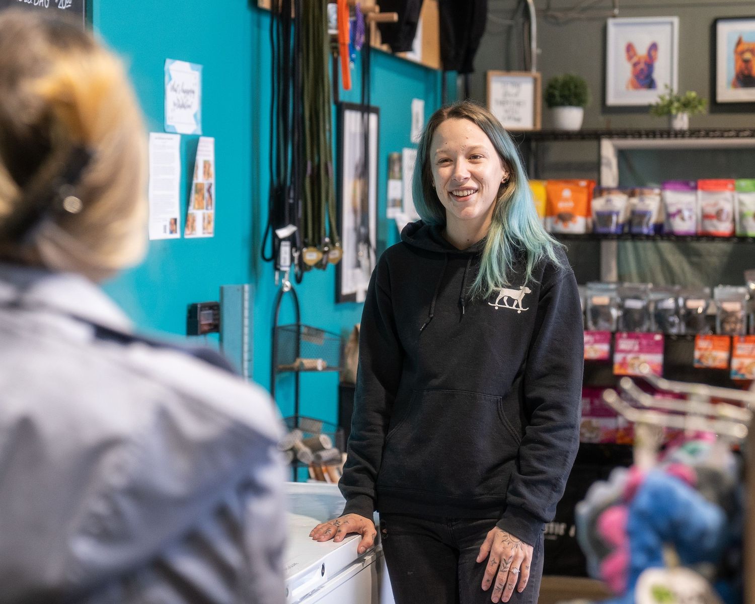 A retail store employee in a black hoodie assists customers at a counter display of merchandise.