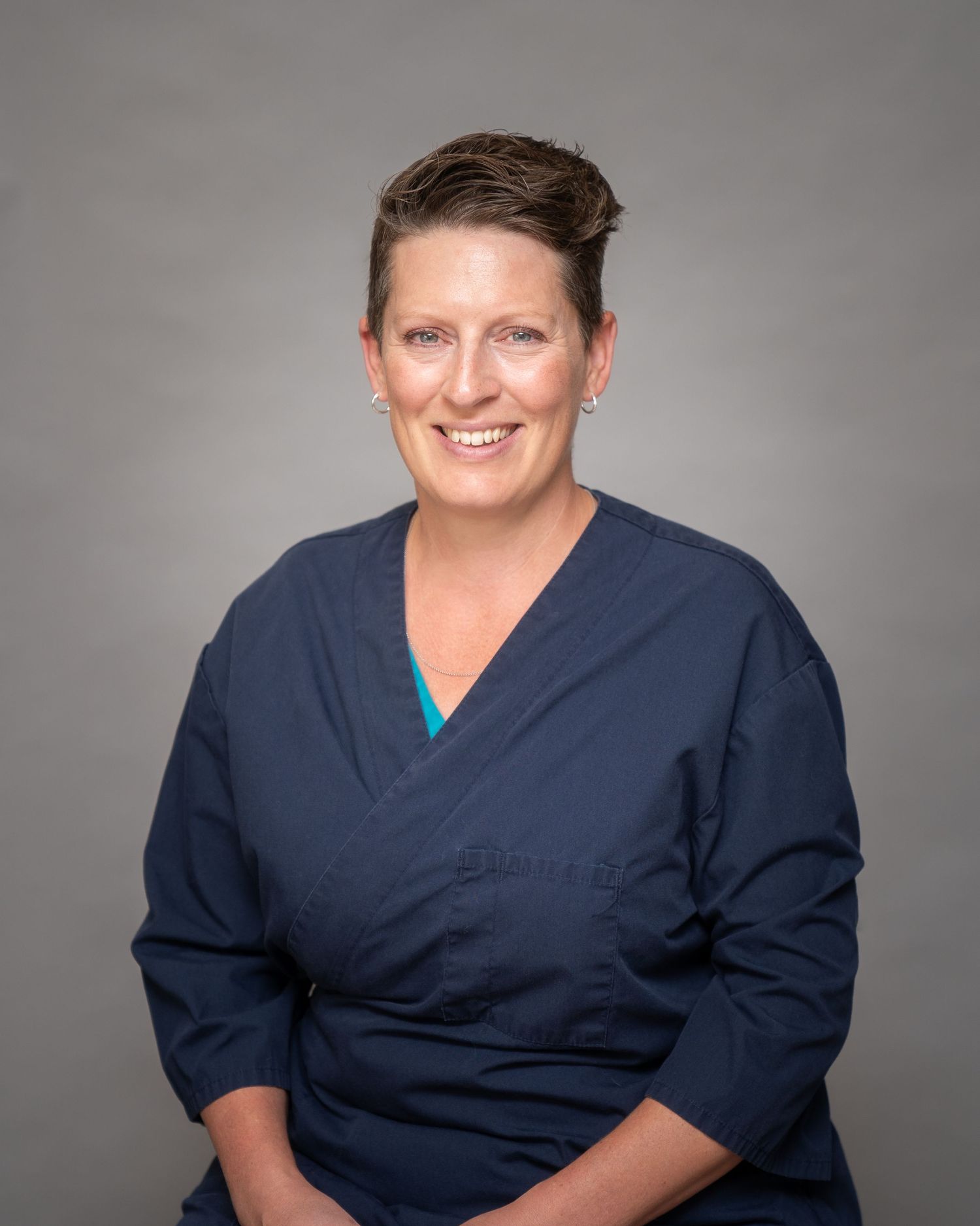 Healthcare professional in navy blue scrubs poses against a gray studio backdrop for a professional headshot series.
