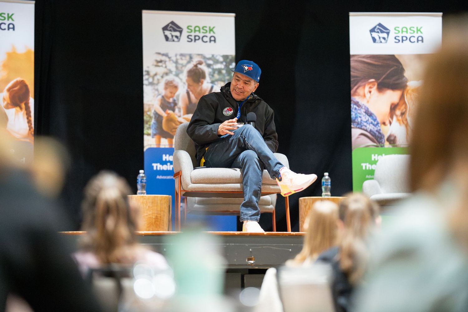 A speaker sits on stage in casual attire during an event with promotional banners displayed behind them.
