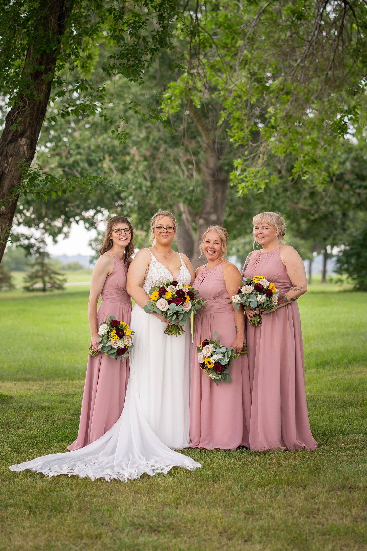 Bridal party in dusty rose dresses poses with bride in white gown on grassy lawn during outdoor wedding photoshoot.