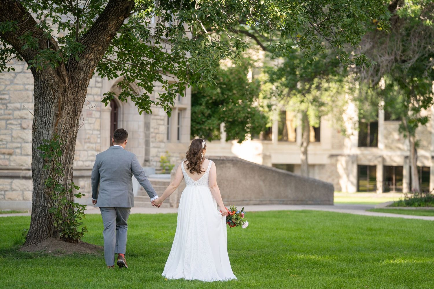 A couple in wedding attire walks hand in hand across a lush green lawn near stone buildings on their special day.