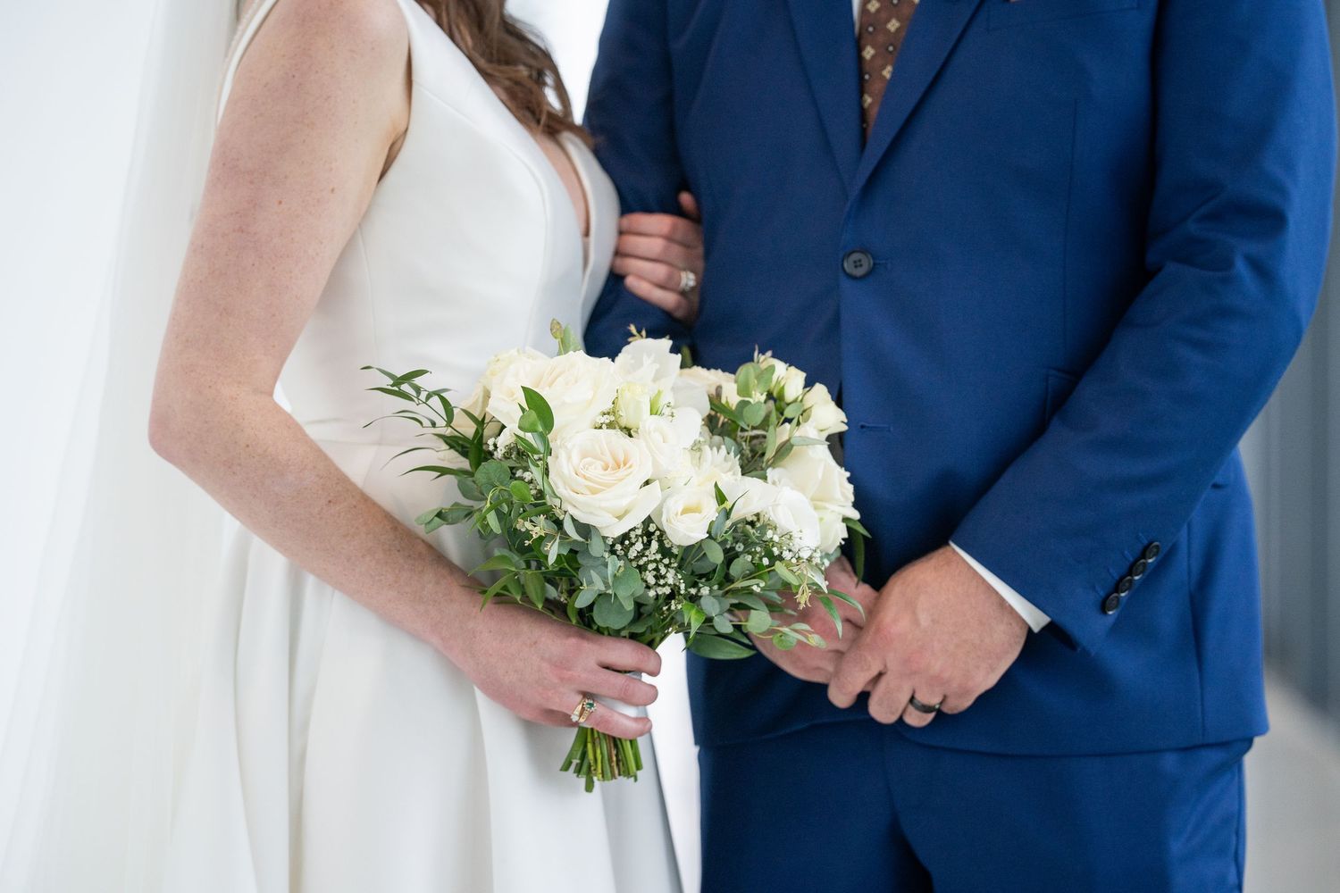 A bride and groom stand together at their wedding ceremony, with the bride holding a white floral bouquet.